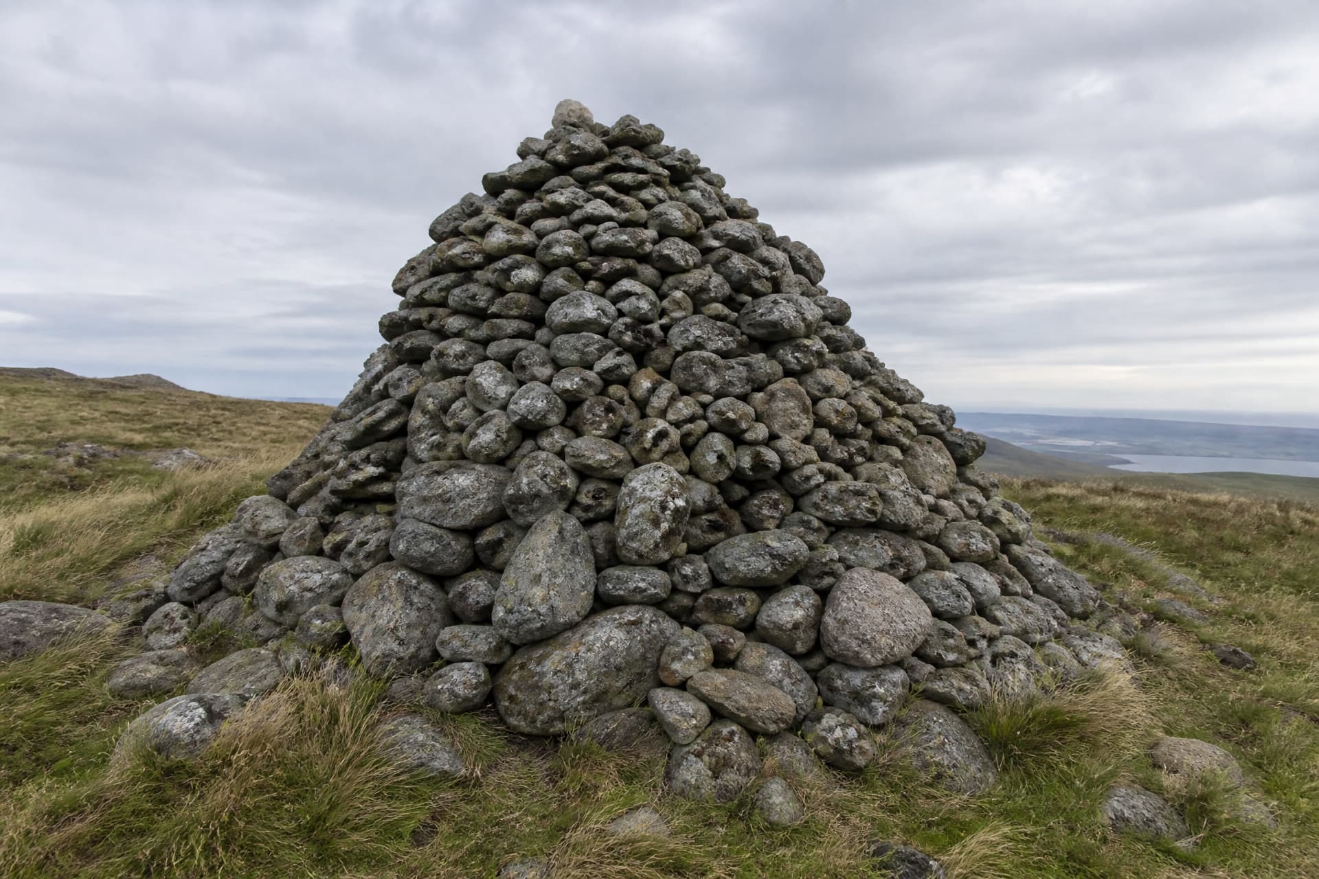 A conical memorial cairn built from several hundred loose lichen-covered field stones, standing taller than a person on an open windswept grass hillside under a heavy overcast sky