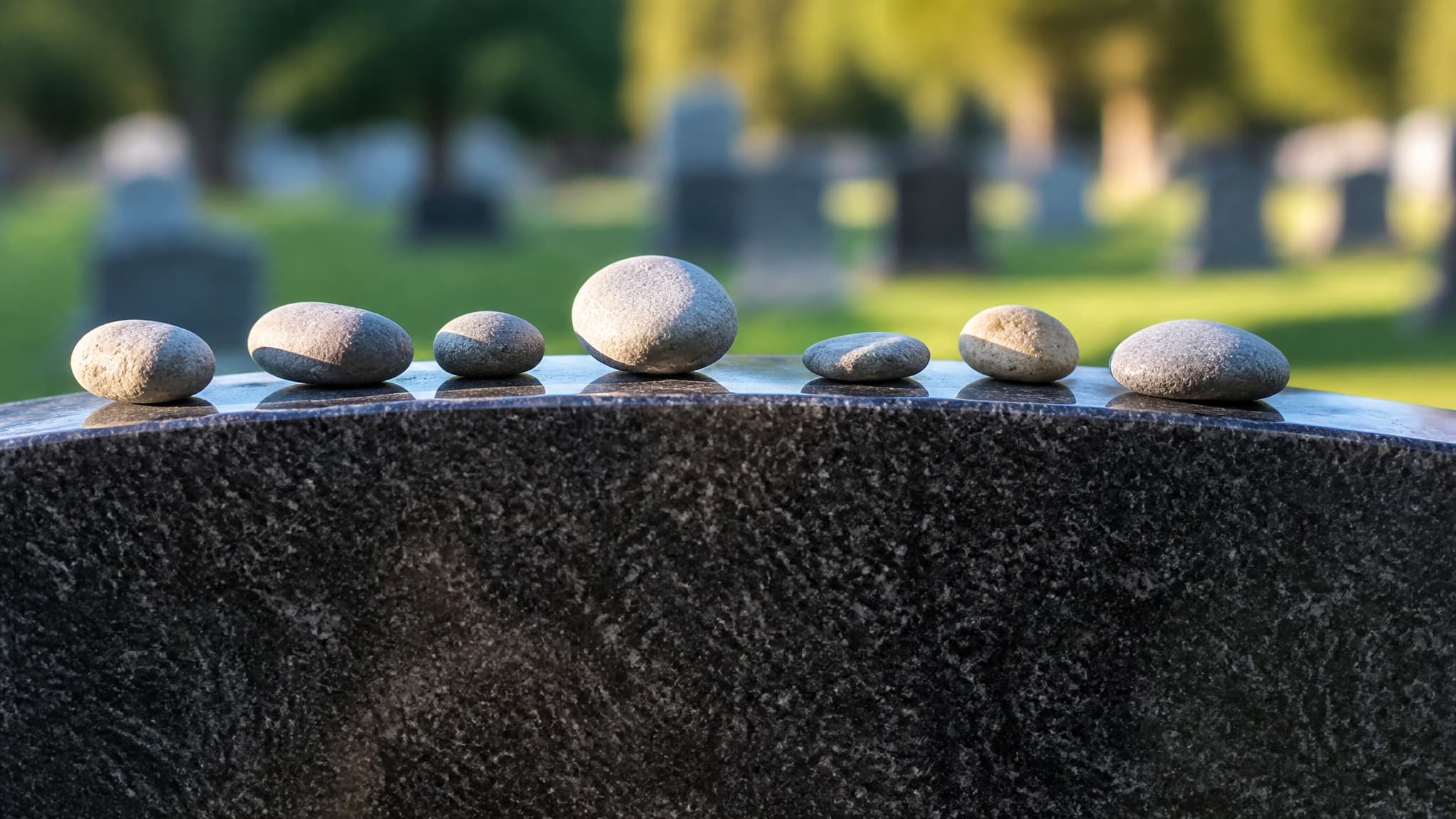 Seven small gray pebbles of different sizes resting along the top edge of a polished granite headstone, photographed close up in late afternoon light with the cemetery lawn blurred behind