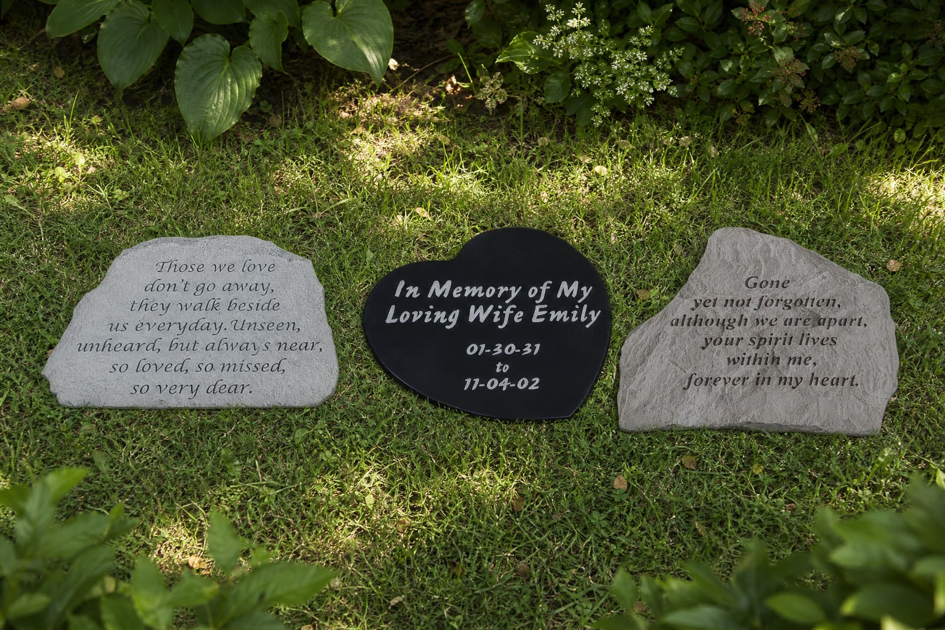 Three engraved memorial stones set among garden planting on a lawn: an irregular gray stone carved with a verse, a black heart-shaped stone with a name and dates, and a second gray stone carved with a longer inscription
