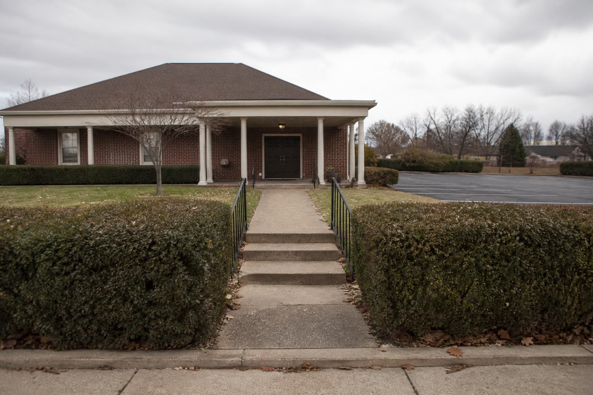 The exterior of an ordinary suburban funeral home on an overcast weekday, low brick with a portico and a short flight of steps, mown grass and a clipped hedge either side of the path, an empty car park in front