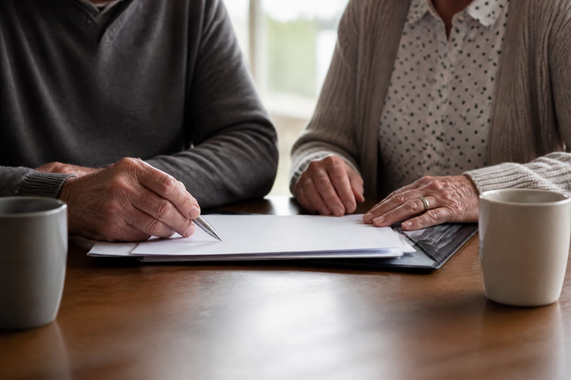 An older couple seated at a dining table seen from chest down, four hands resting on and around a folder of paperwork between them, one hand holding a pen, mugs at the edge of the table