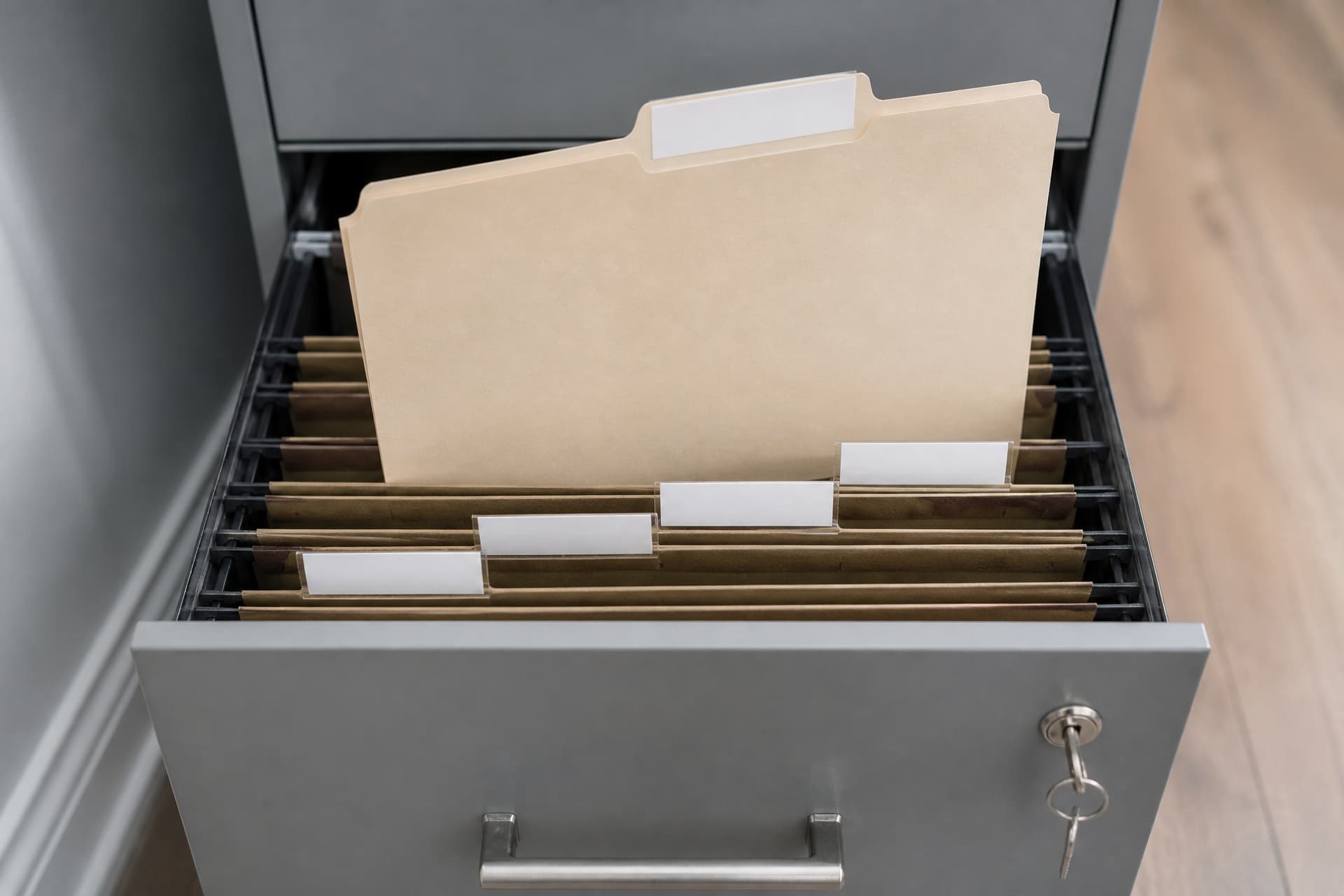 An open domestic filing drawer seen from above, hanging files with blank tabs, one folder lifted clear of the others and a small key in the lock of the cabinet