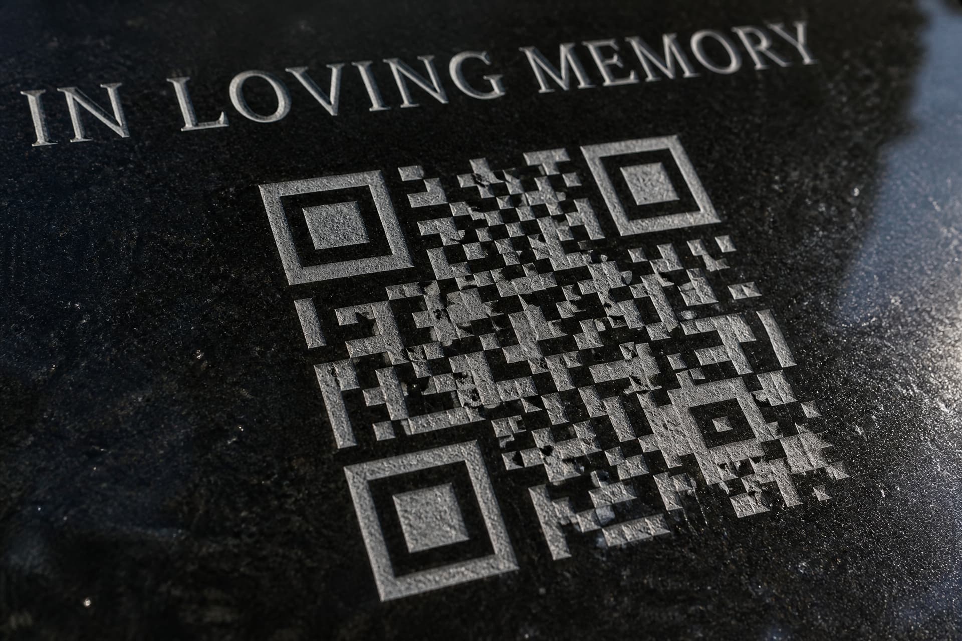 A macro view of a QR code sandblasted directly into polished black granite, the cut squares pale gray against the mirror-black stone with the carved capitals IN LOVING MEMORY partly visible above