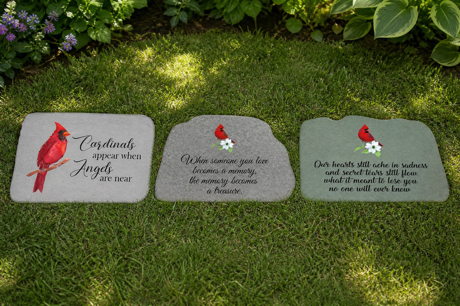 Three engraved memorial garden stones lying flat on the grass beside a planting bed, each carrying a short cardinal line