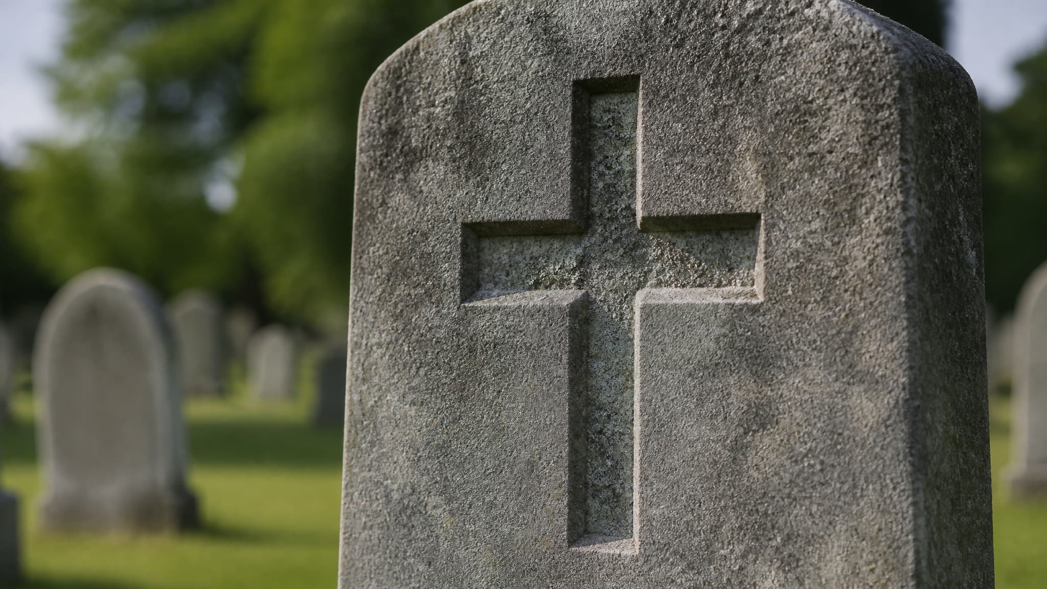 A close view of a carved stone cross on a weathered granite headstone in raking afternoon light, lichen in the recesses and a blurred churchyard behind
