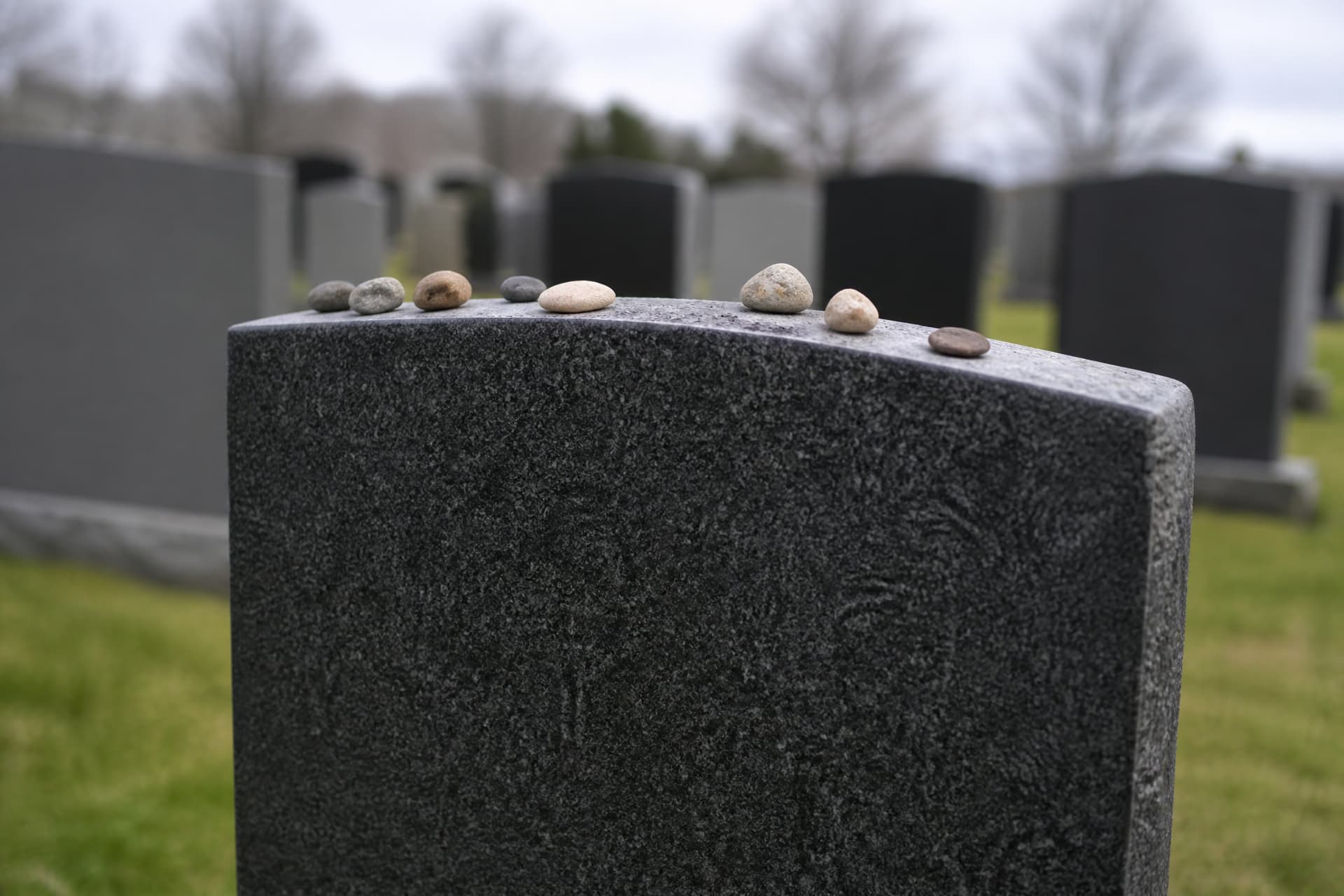Several small smooth pebbles resting along the top edge of an upright dark granite headstone in a Jewish cemetery, more stones out of focus behind, the face of the stone turned away so no lettering shows