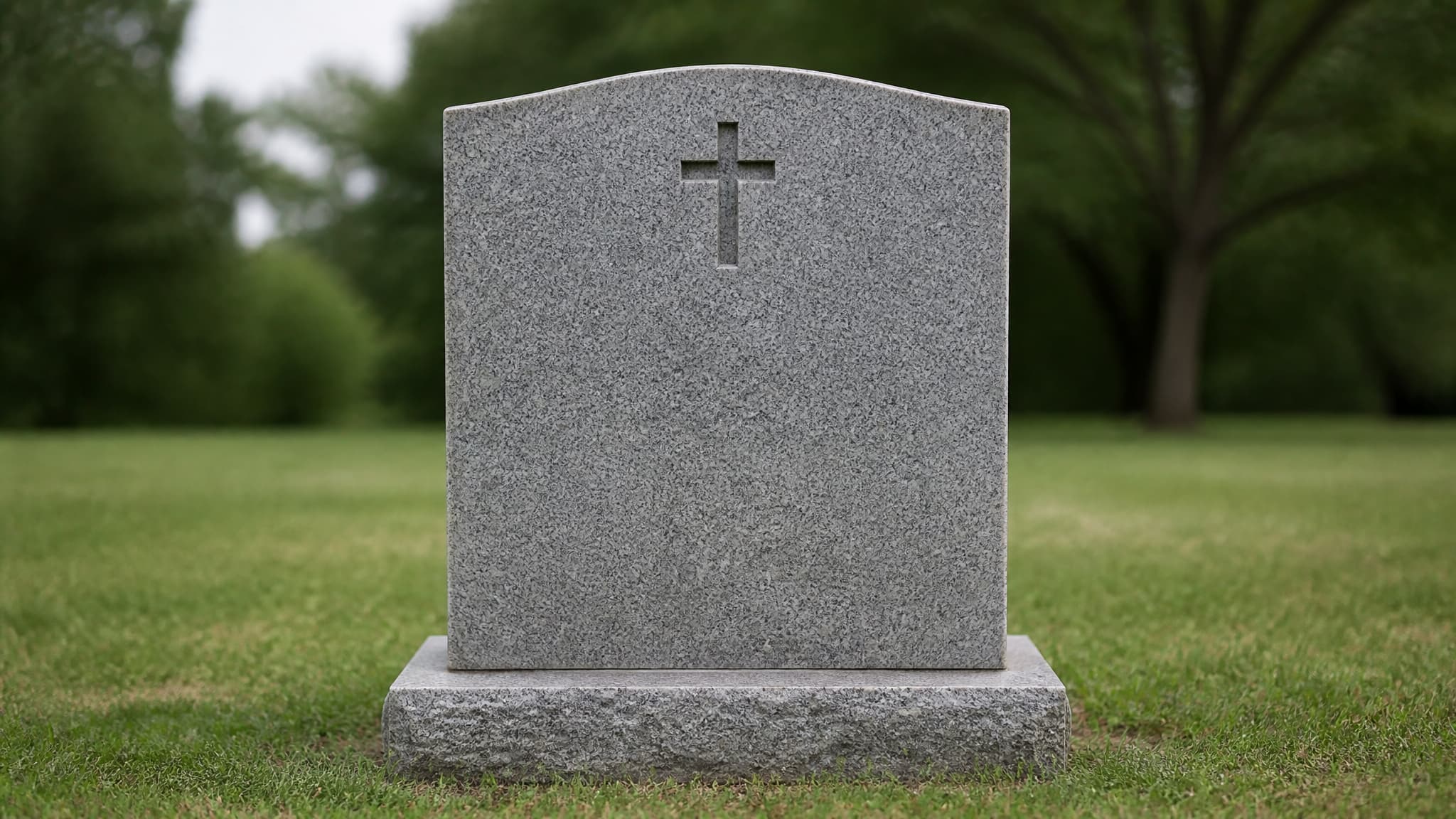 A granite headstone with a simple carved cross, photographed straight on in soft daylight with mown grass around it and out-of-focus trees behind