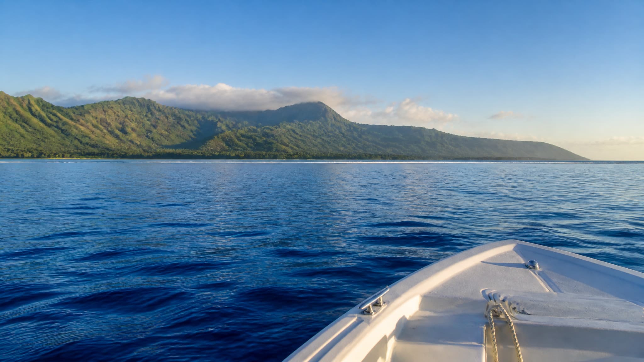 A view from a small boat on calm open ocean off a Hawaiian island in early morning light, the green volcanic coastline low on the horizon behind, deep blue water in the foreground and a faint line of reef visible closer to shore