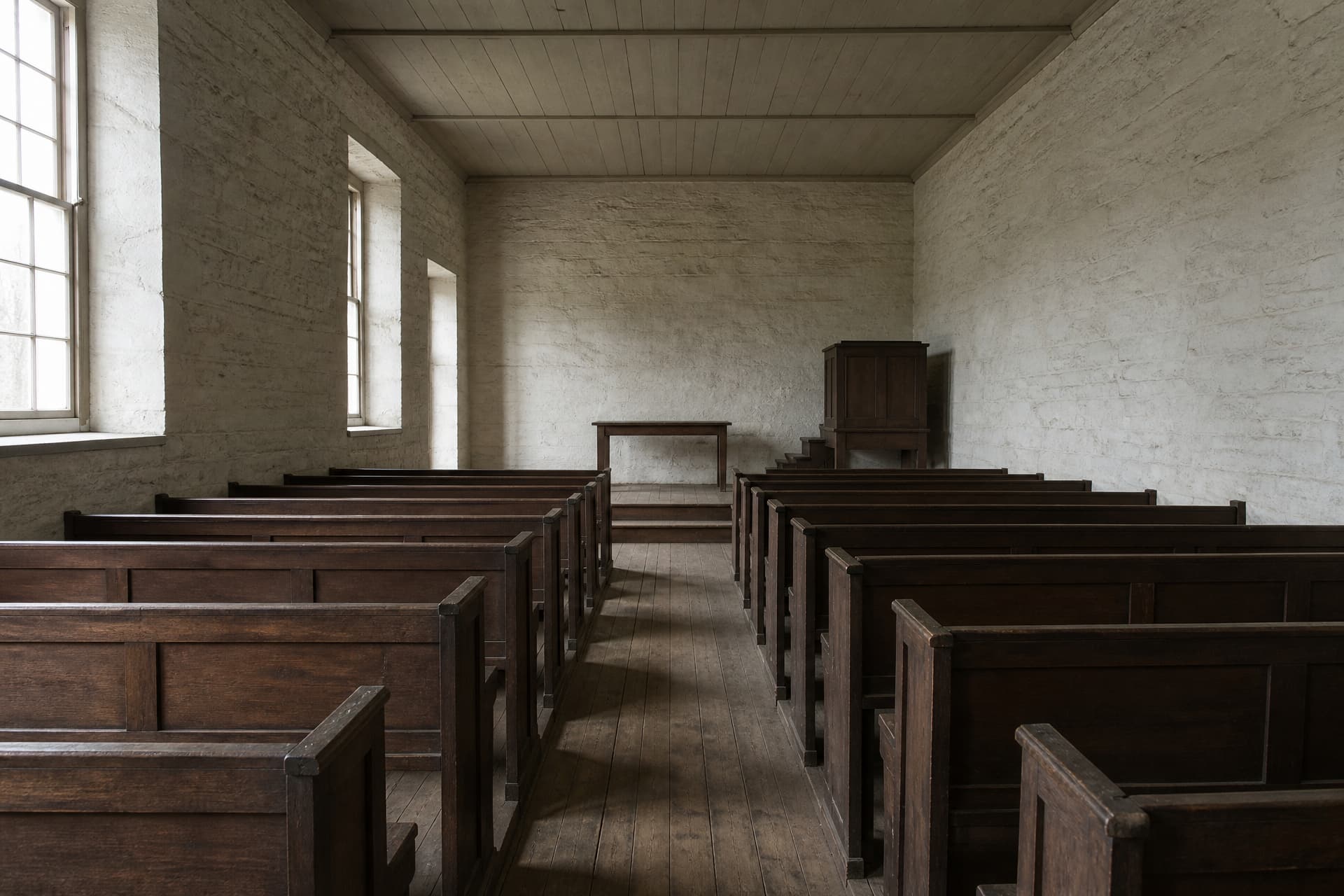The inside of a plain Presbyterian kirk seen from the back of the center aisle, with lime-washed stone walls, clear glass windows, dark wood pews, a bare wooden communion table on a low platform and a simple raised pulpit beside it