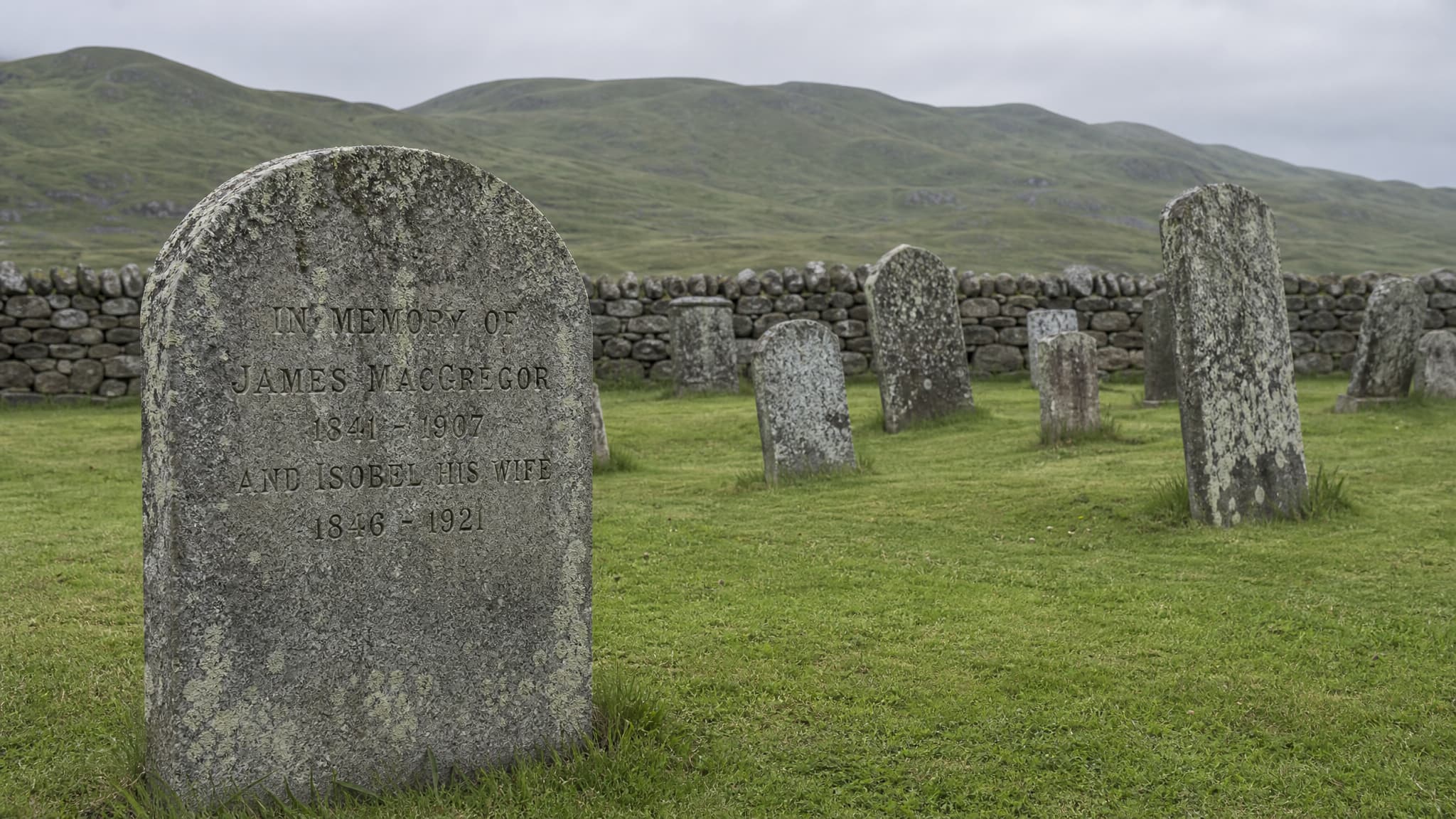 A Scottish kirkyard on an overcast morning โ a lichen-covered granite headstone reading In Memory of James MacGregor in the foreground, older leaning stones behind it, a low drystone wall and green hills beyond