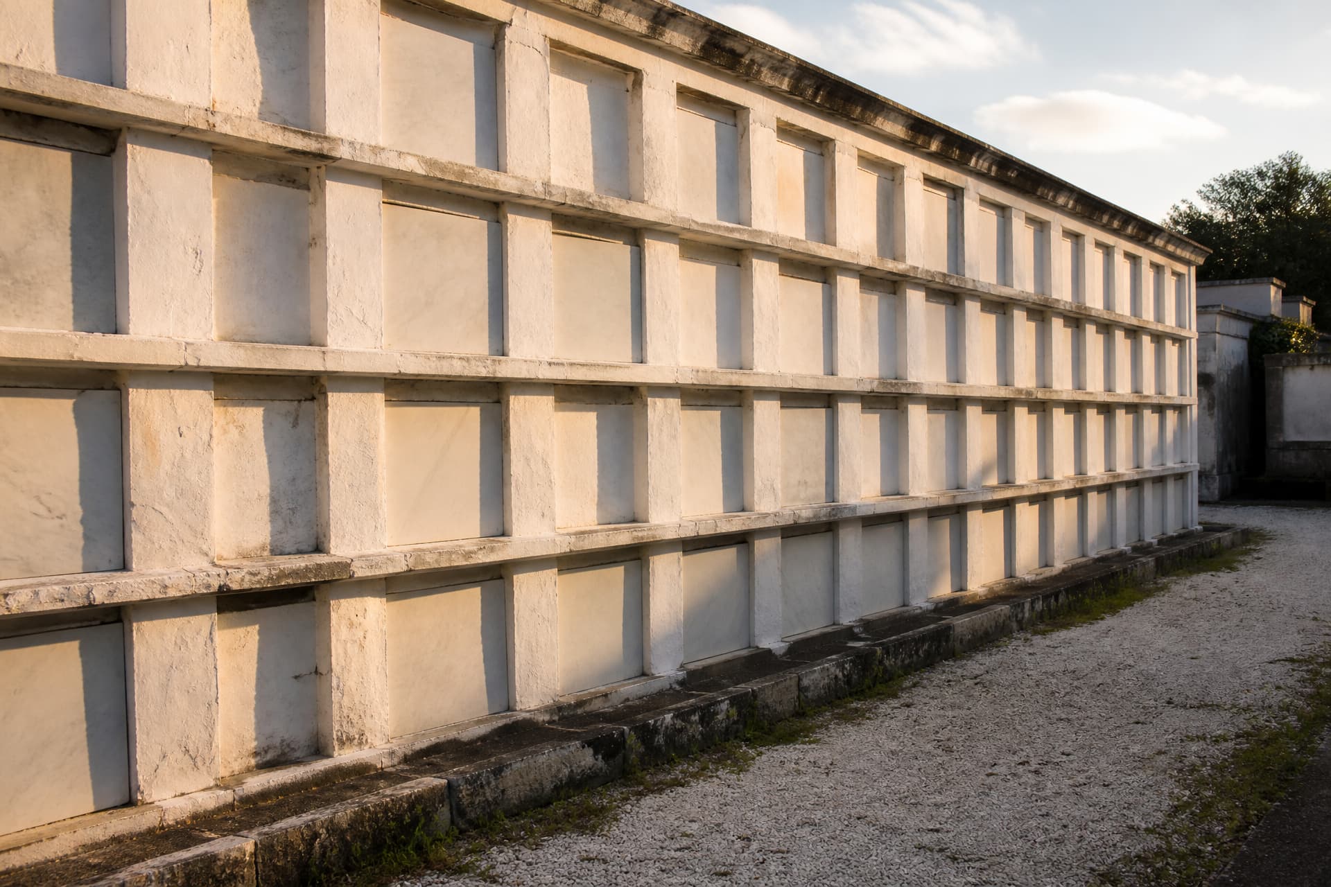 A weathered whitewashed wall of stacked above-ground cemetery vaults with closed stone tablet fronts, photographed in warm late afternoon light
