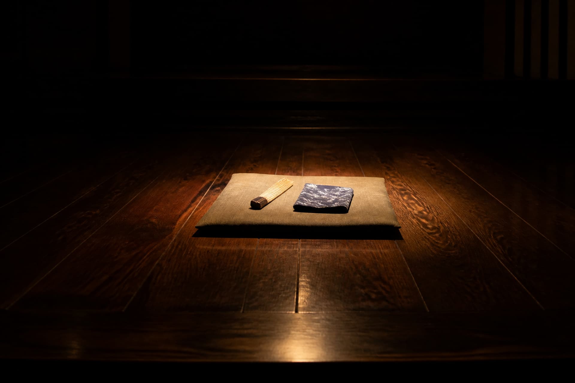 A dark polished wooden stage floor lit by a single warm overhead light, with a flat cushion at the center holding a closed folding paper fan and a folded cloth, the rest of the room in deep shadow