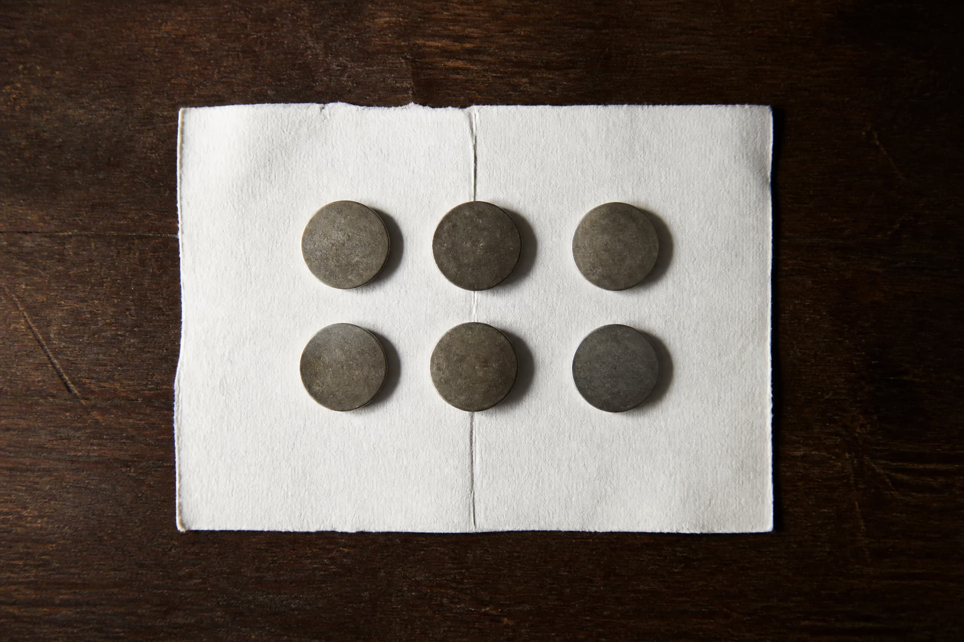 Overhead view of six small plain metal coins laid in two rows on a folded sheet of white paper on a dark wood surface, lit softly from the left