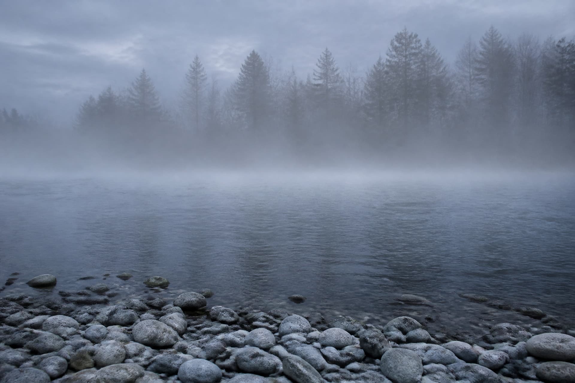 A wide shallow river at first light with low mist lying across the surface, smooth pale stones along the near bank, and dark trees barely visible on the far side in cold gray-blue light