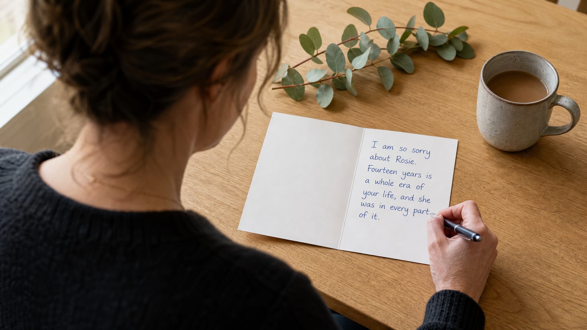 Seen over her shoulder, a woman writing a sympathy message in a card lying open on a kitchen table