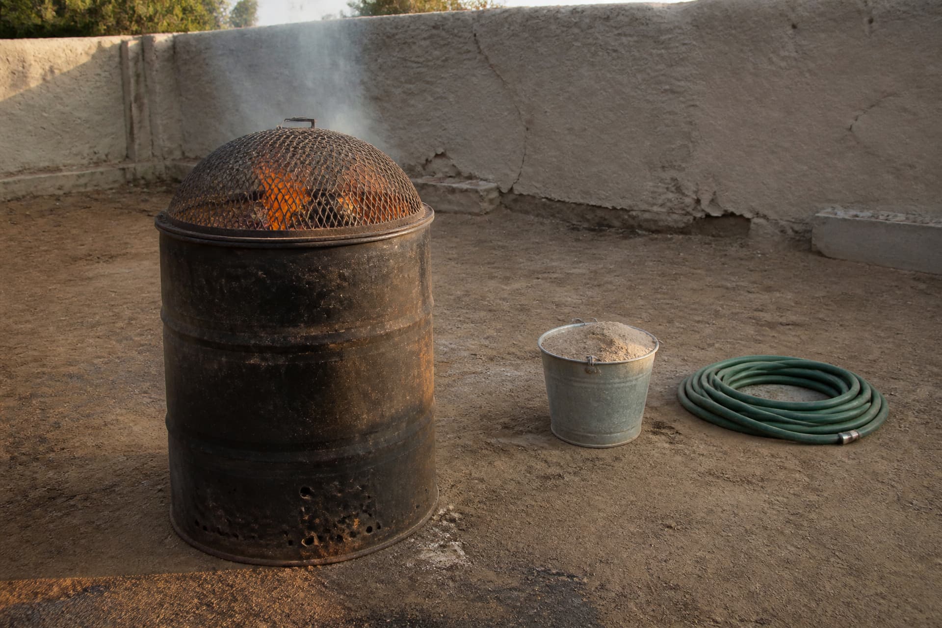 A blackened metal burning drum with a mesh lid standing on bare ground in a courtyard, low flames visible through the mesh, a metal bucket of sand and a coiled hose on the ground beside it