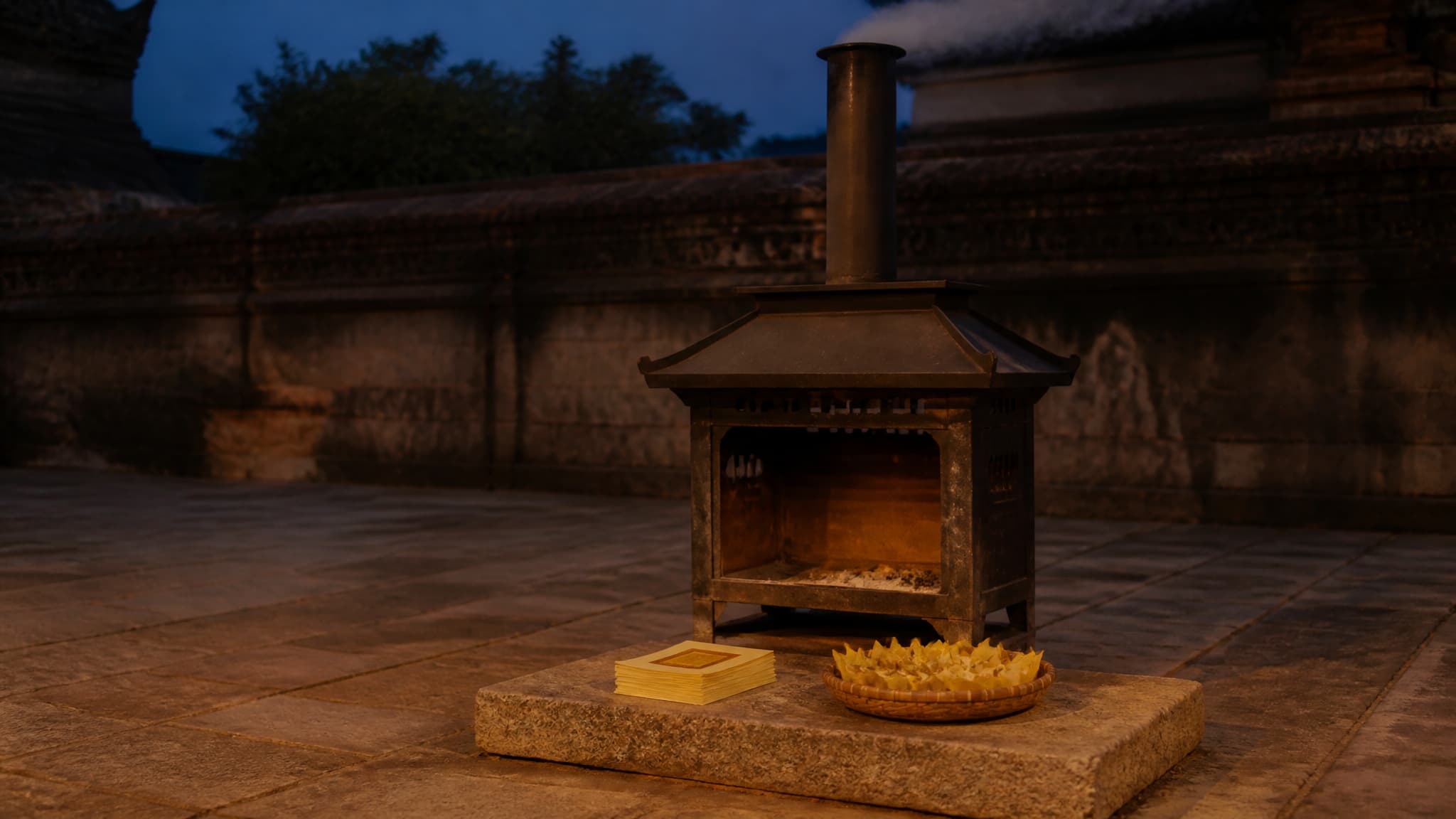 A pagoda-roofed metal burner with a chimney standing in a stone temple courtyard at dusk, a stack of gold foil paper squares and a basket of folded paper ingots waiting on the stone step in front of it