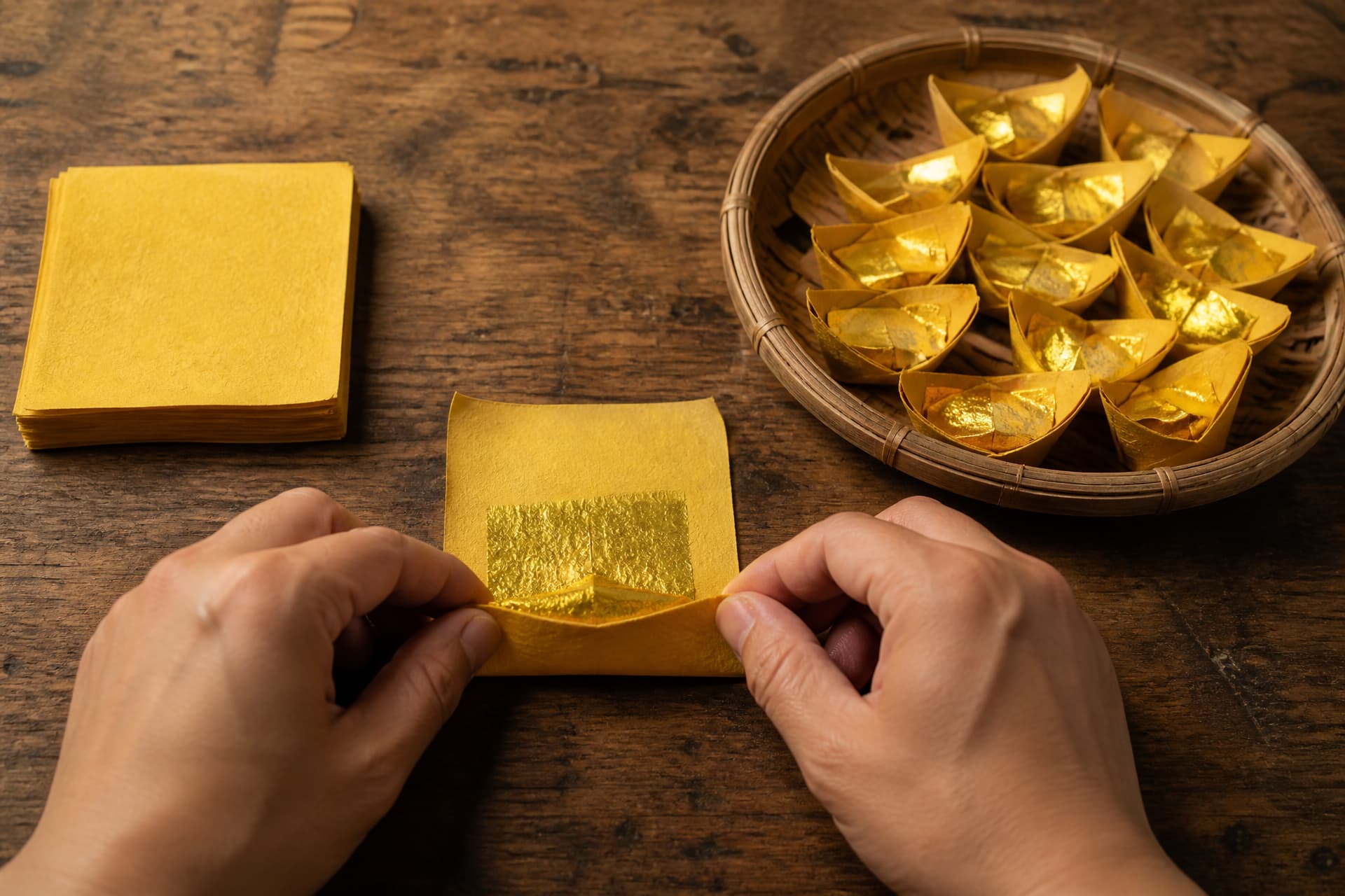 A pair of hands folding a square of yellow paper with a gold foil center into a boat-shaped ingot, with a low basket of finished ingots beside them and a stack of unfolded squares on the other side