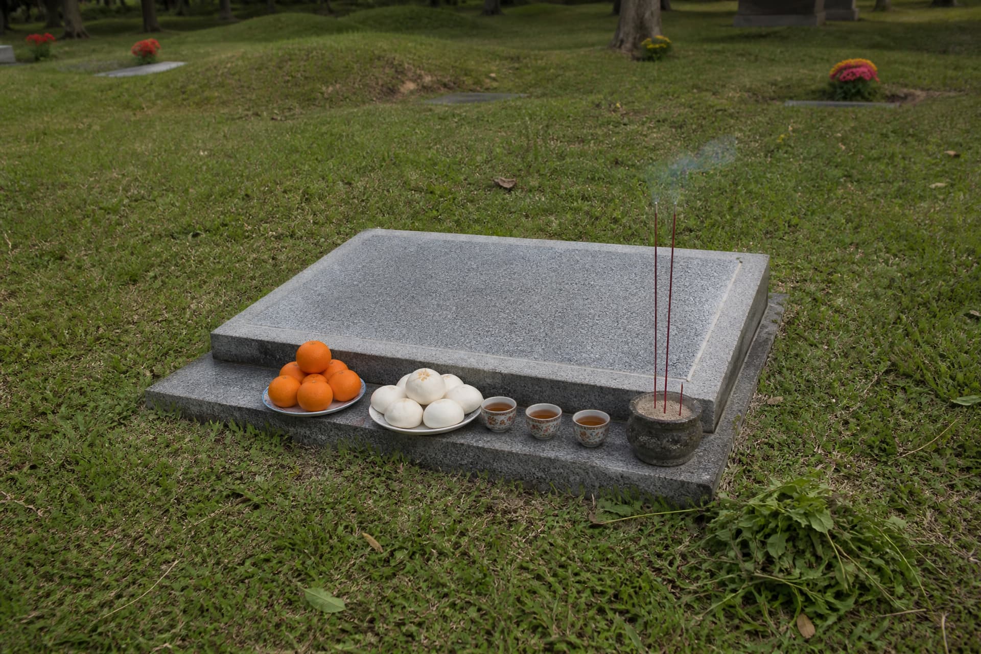A swept granite grave marker with fresh weeds pulled and stacked to one side, a plate of oranges and steamed buns and three cups of tea set on the stone ledge, and three sticks of incense standing upright in a small pot of sand