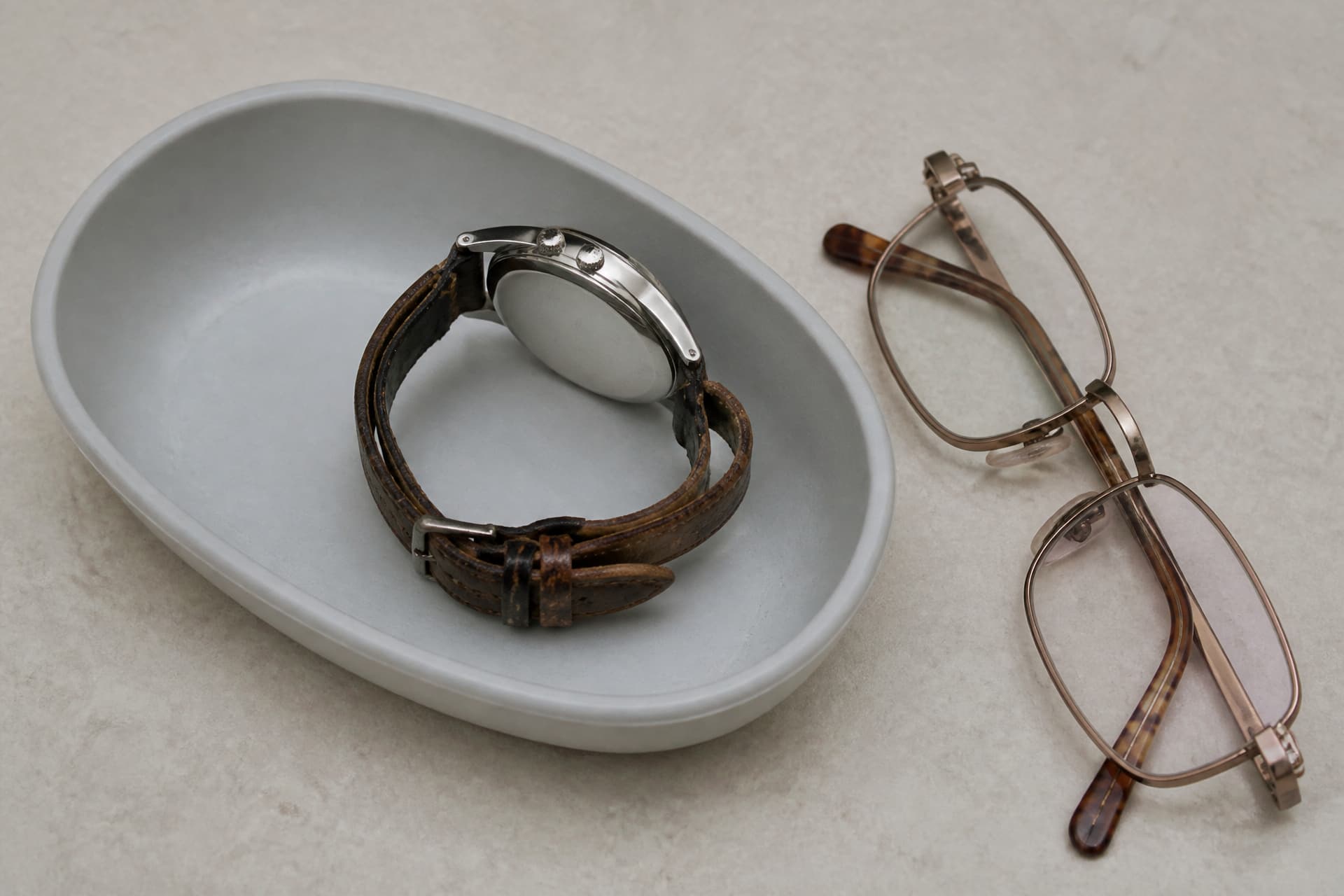 A worn leather-strapped wristwatch lying in a small plastic dish on a hospital nightstand beside a folded pair of glasses