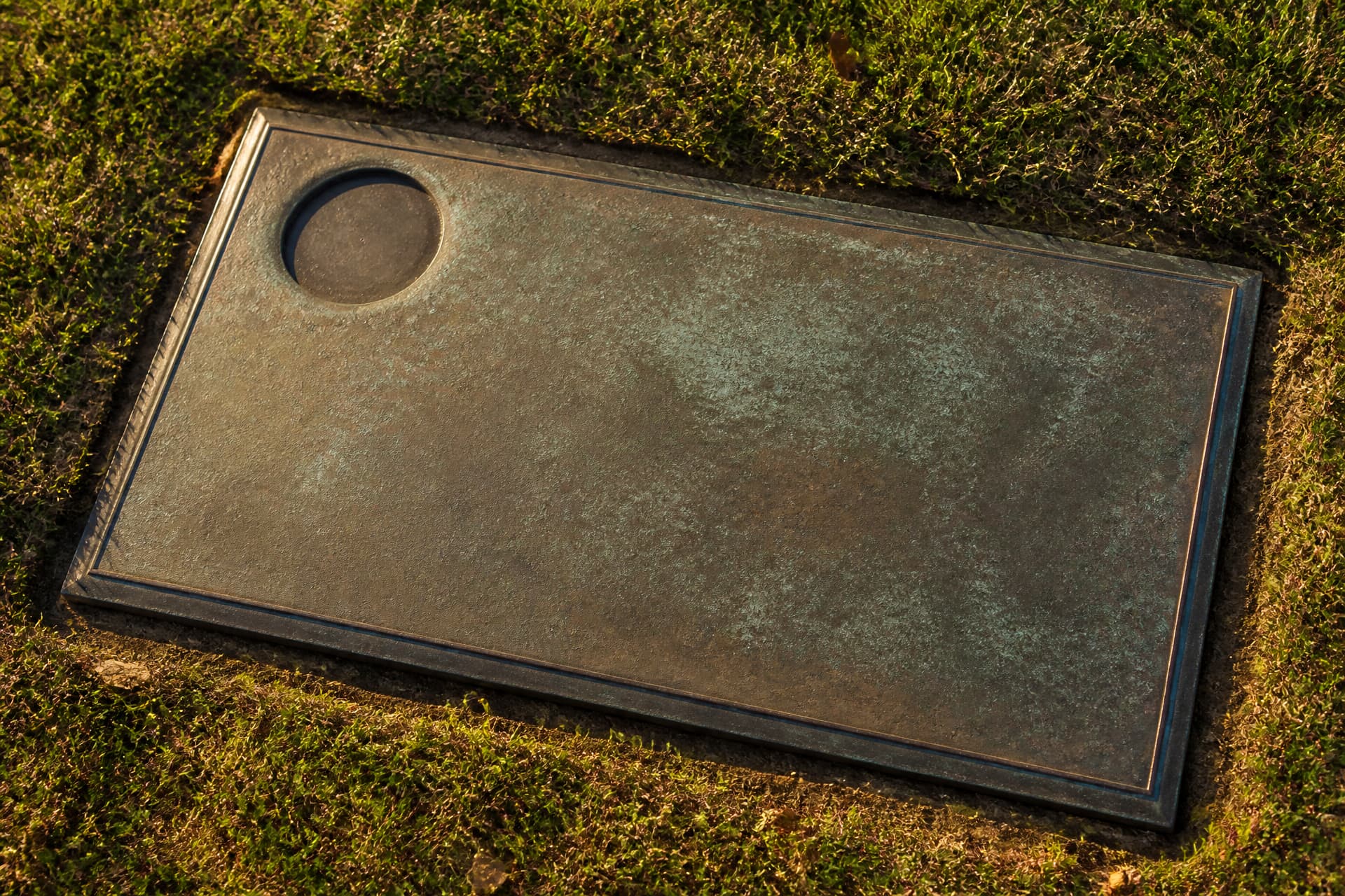 A flat bronze grave marker set flush in cemetery grass with a plain empty circular recess in one corner, weathered patina, low afternoon light
