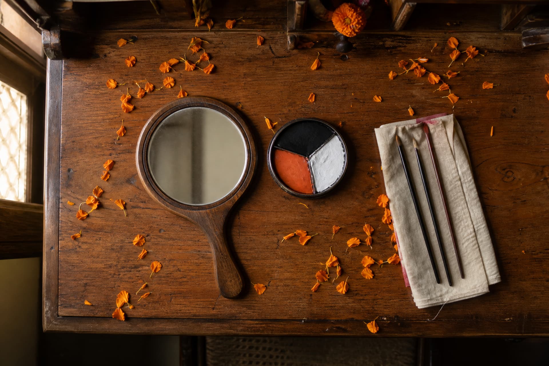 A dressing table with a hand mirror lying flat, an open tin of face paints in orange white and black, three fine brushes, and a scatter of orange marigold petals across the wood