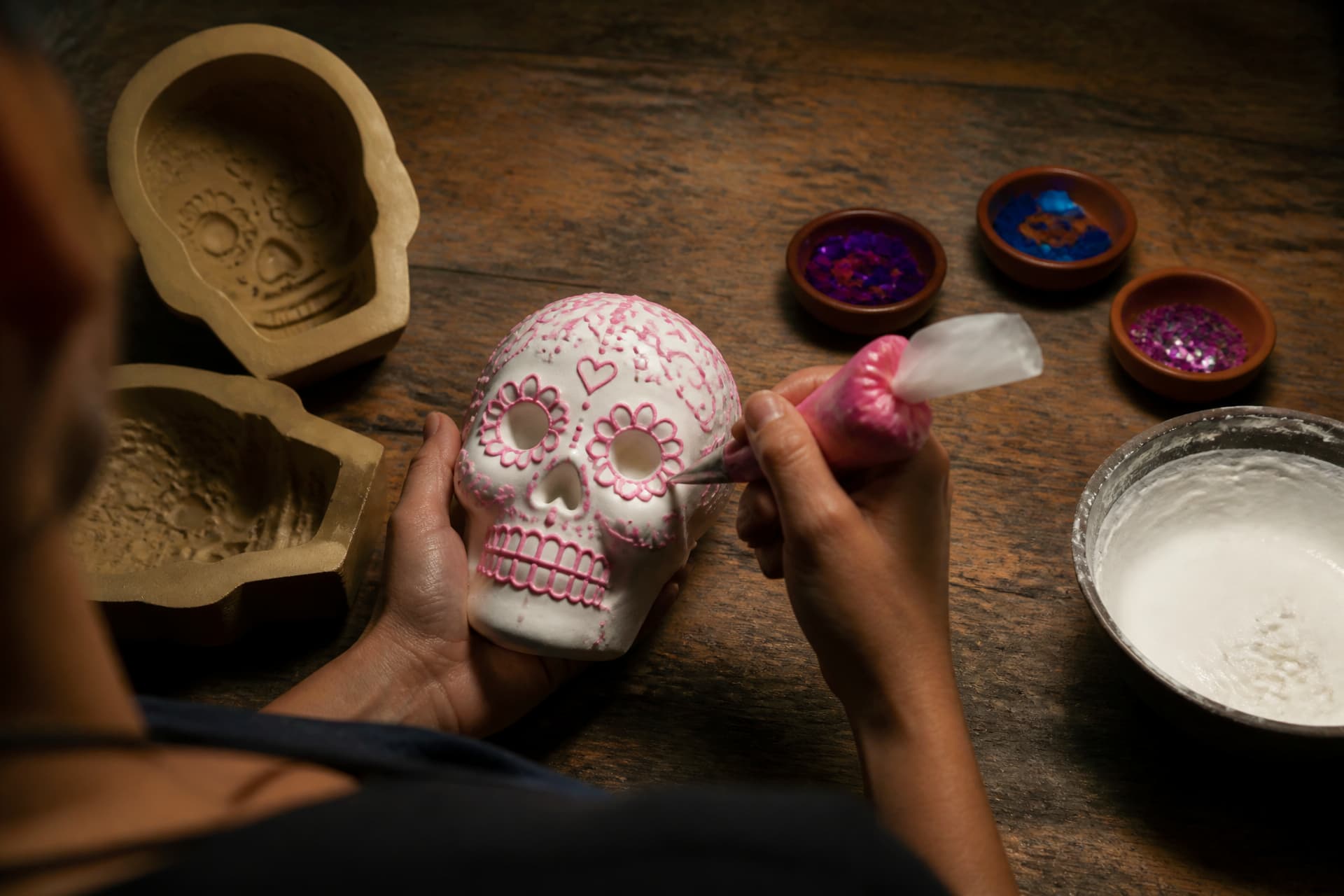 A pair of hands piping bright pink icing onto a plain white sugar skull on a scarred wooden workbench, with two-part clay molds, a bowl of white icing and small dishes of colored foil and sequins arranged around it