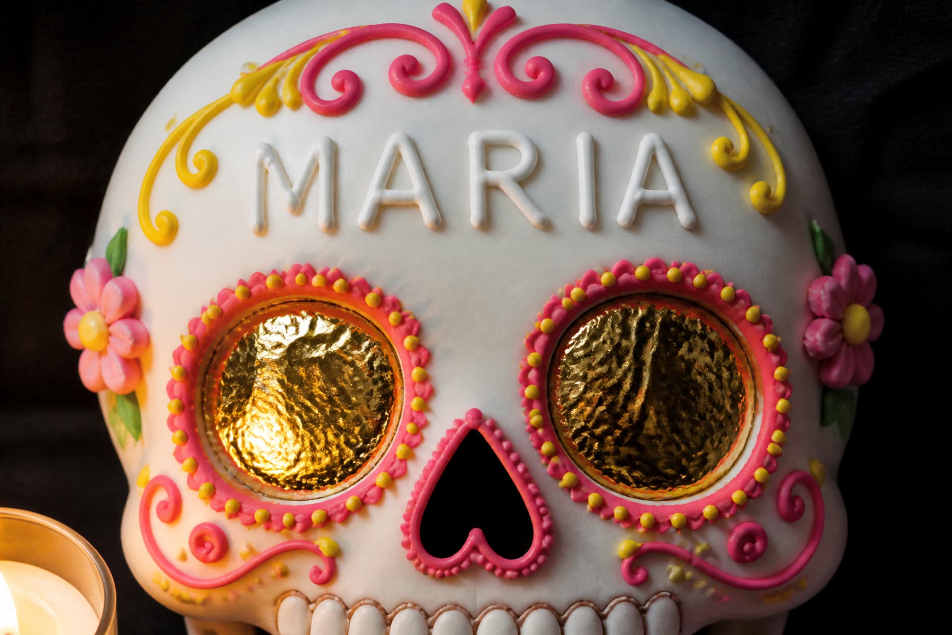 A single large white sugar skull photographed close up on a dark cloth, with the name MARIA piped across its forehead in white icing above bright pink and yellow scrollwork and round foil eyes