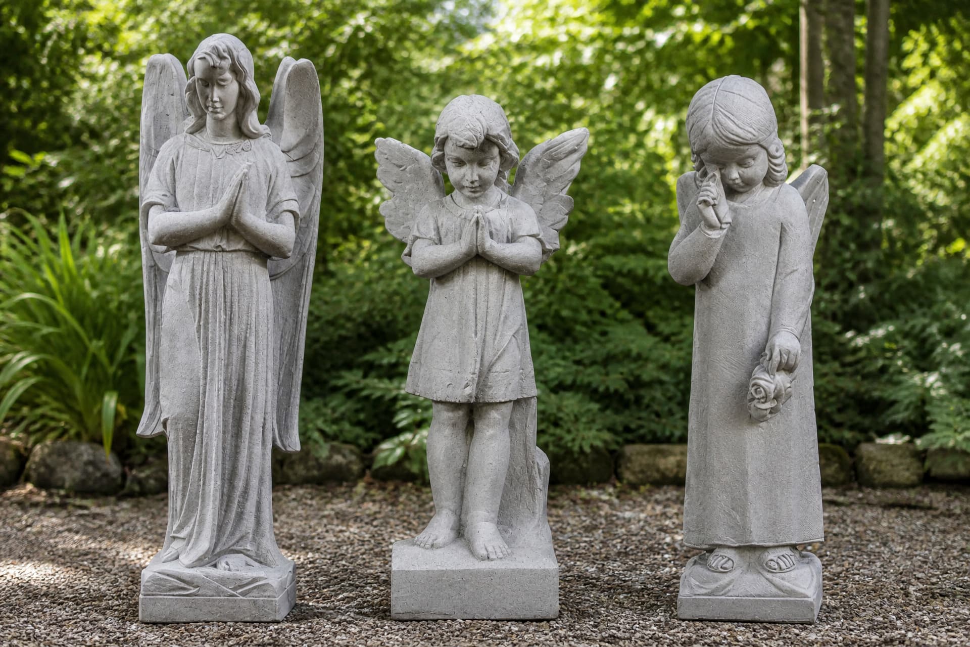 Three carved stone angel statues standing in a row on a gravel path in a green garden, two praying and one with a hand raised to her face