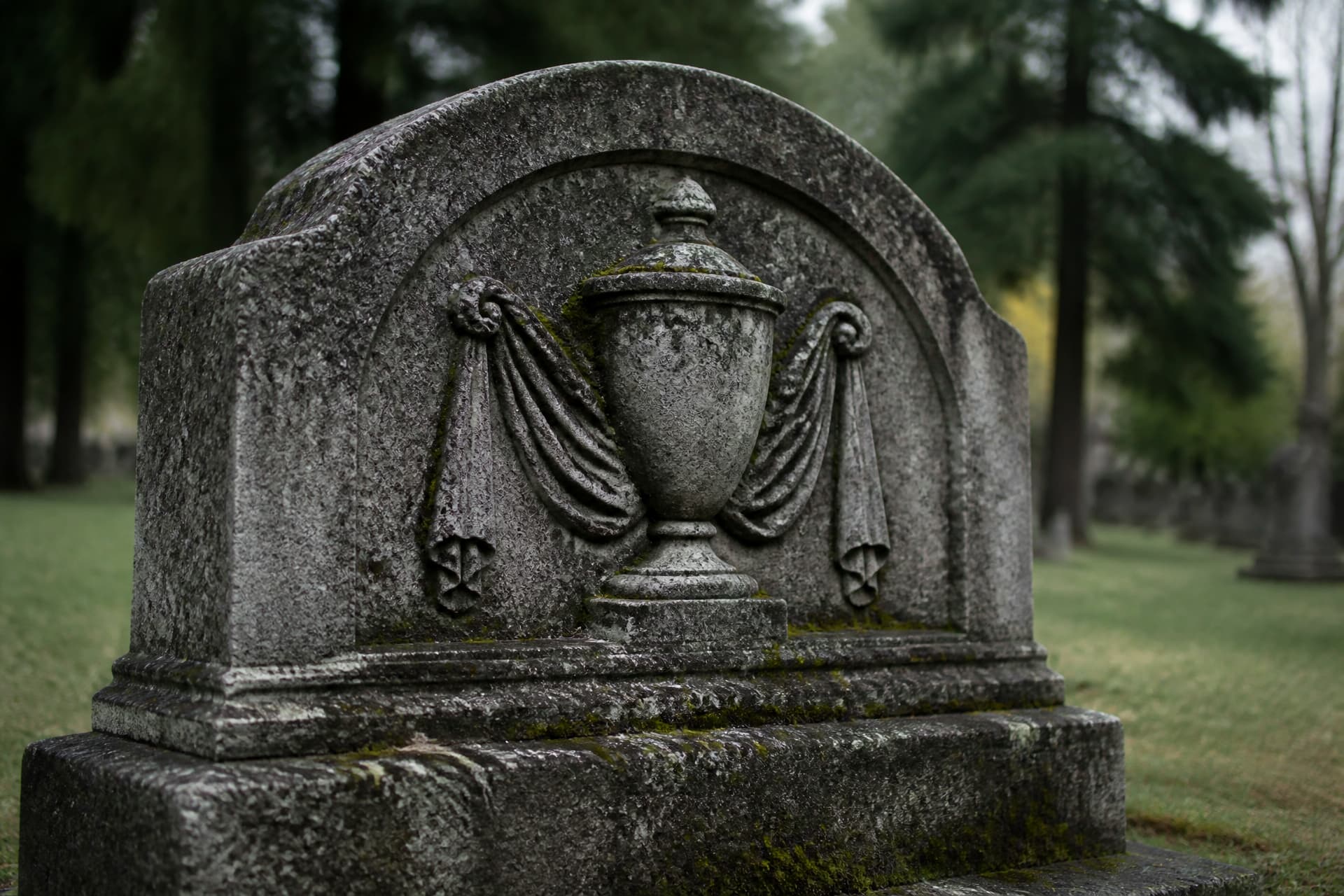A weathered granite memorial carved with a draped urn motif, photographed from a low angle in an old cemetery under soft daylight