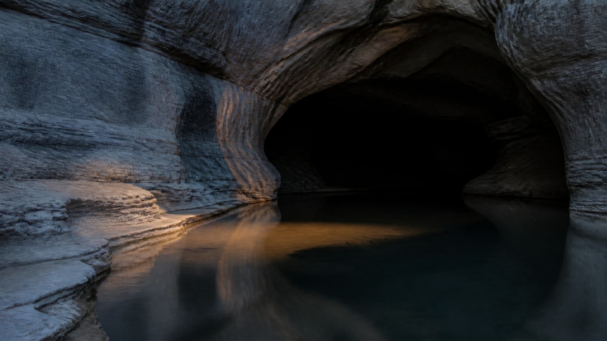 A dark still pool of water at the mouth of a worn limestone cave at dusk, one shaft of low warm light falling across the surface, deep shadow beyond the opening