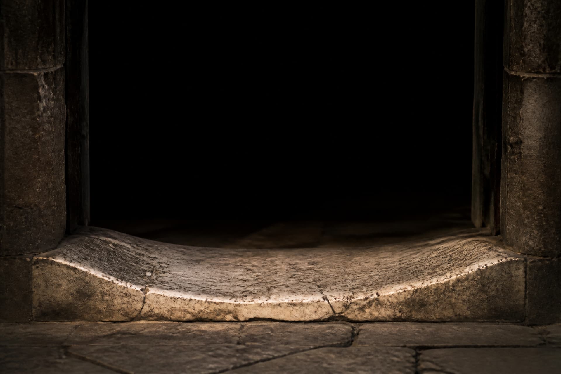 A heavy weathered marble threshold stone set into a dark stone doorway, worn smooth in the center by centuries of footsteps, warm light falling on the stone and total darkness beyond the opening