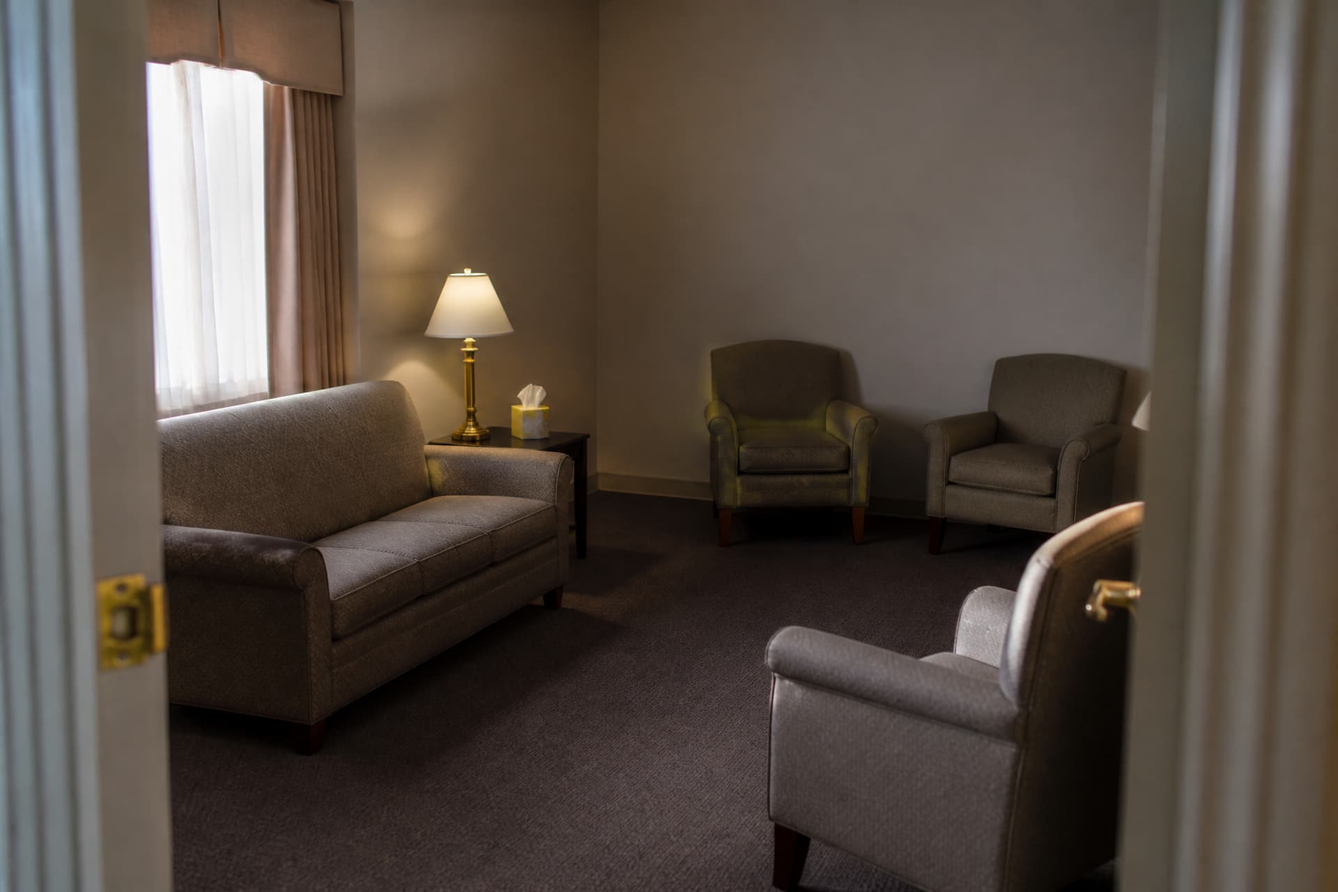 An empty funeral home visitation room seen from the doorway, upholstered armchairs and a sofa in loose groups rather than rows, a side table with a lamp and a box of tissues, half-drawn curtains