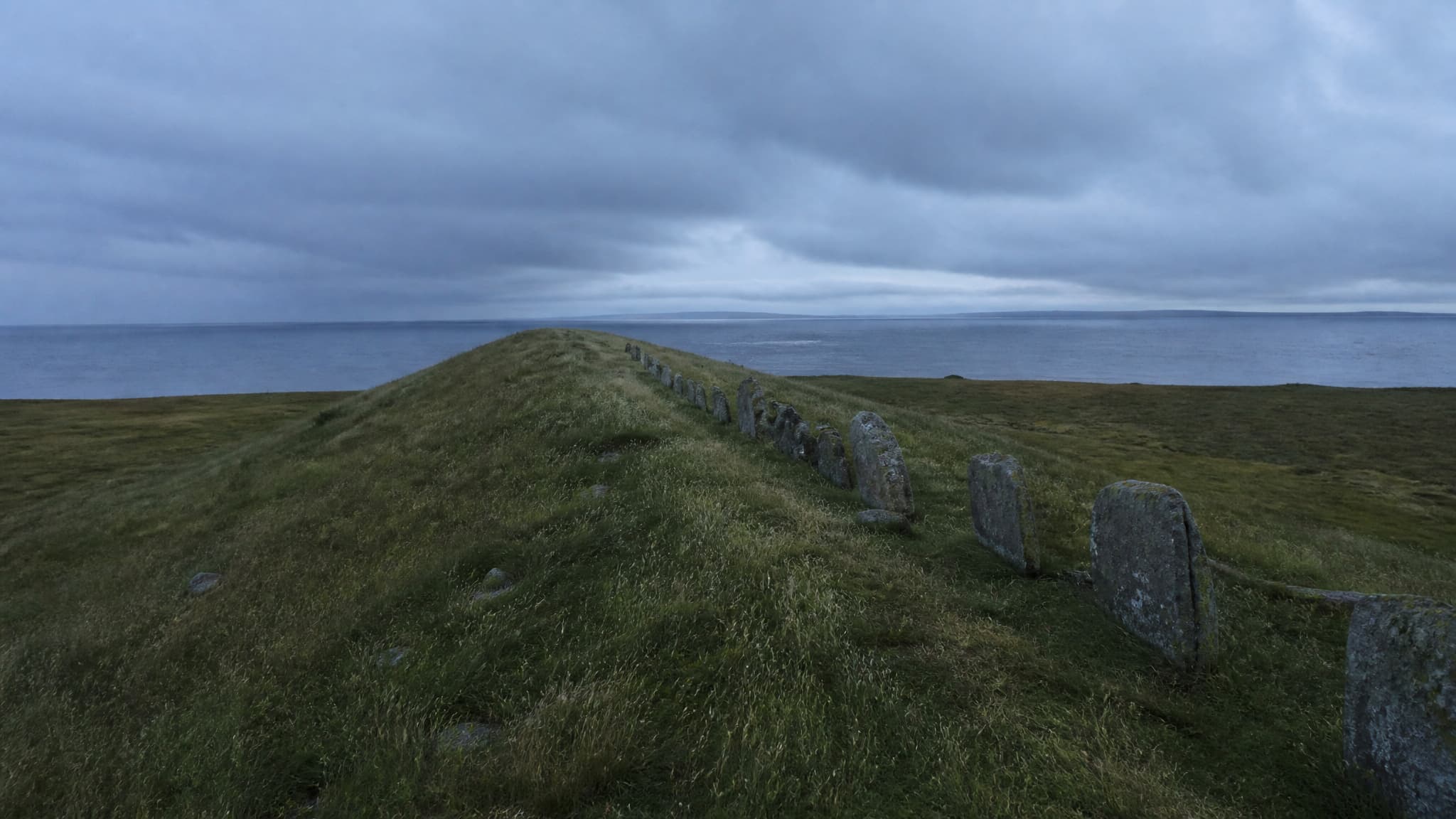 A grass-covered Norse burial mound at dusk on a northern coastline, with standing stones at its edge and gray sea behind it