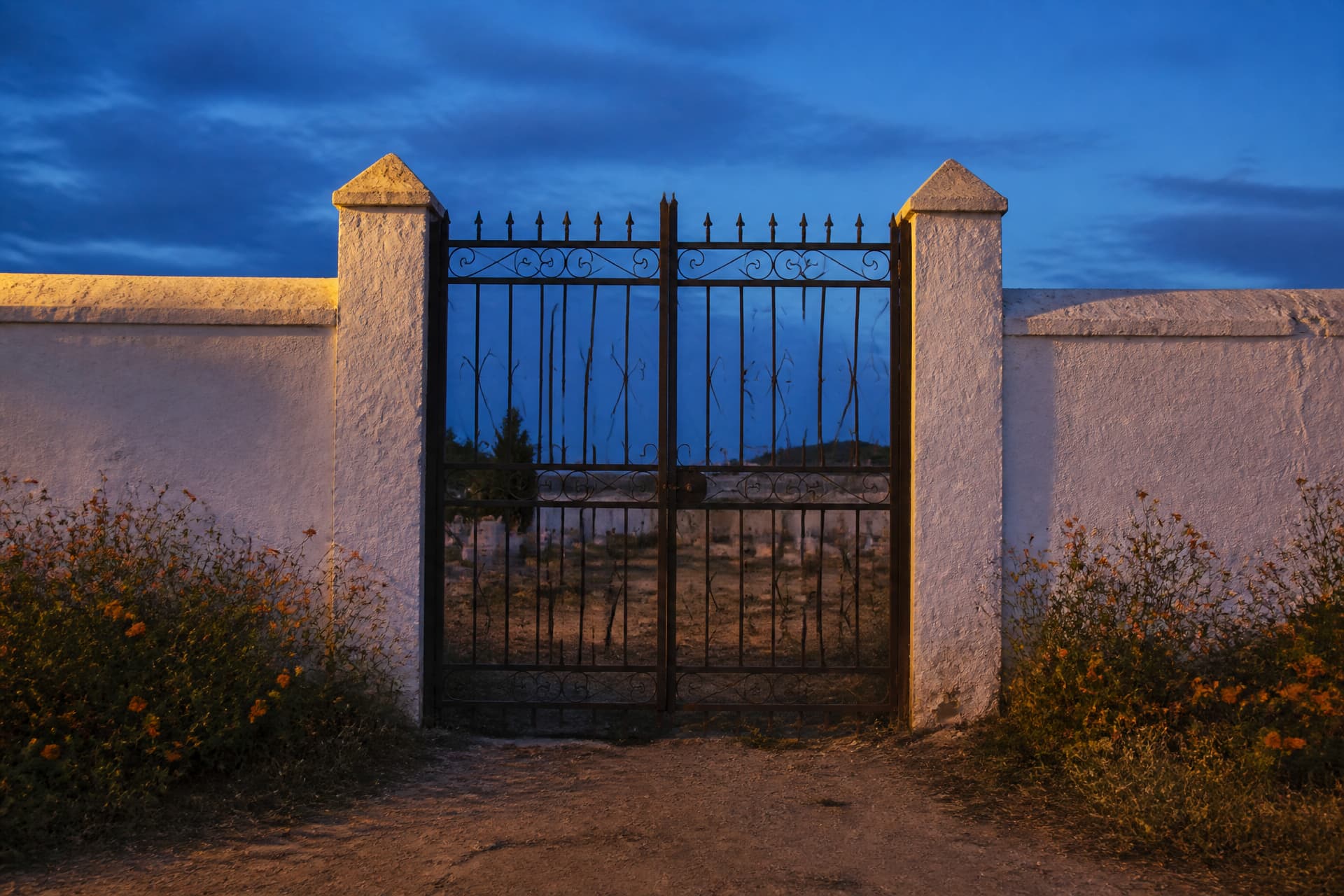 A closed wrought-iron cemetery gate set into a whitewashed wall at dusk, with marigolds growing wild along the base of the wall under a deep blue sky