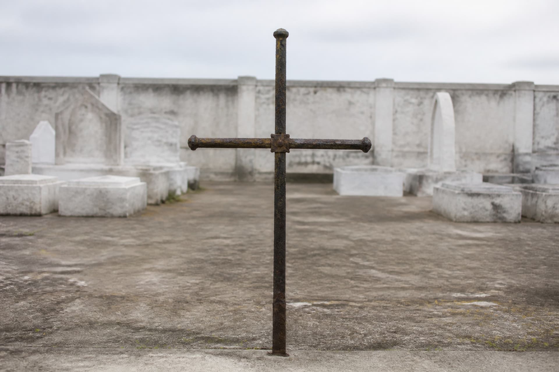 A weathered iron cross standing alone at the center of a walled cemetery yard under flat overcast light, plaster tombs out of focus behind it