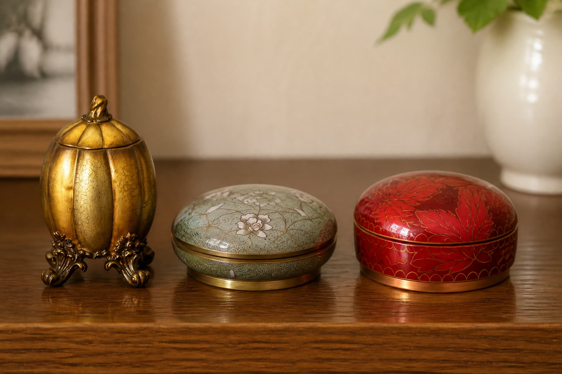 A gilded footed keepsake urn, a low green cloisonne box with a domed lid, and a deep red cloisonne box painted with maple leaves, on a wooden sideboard