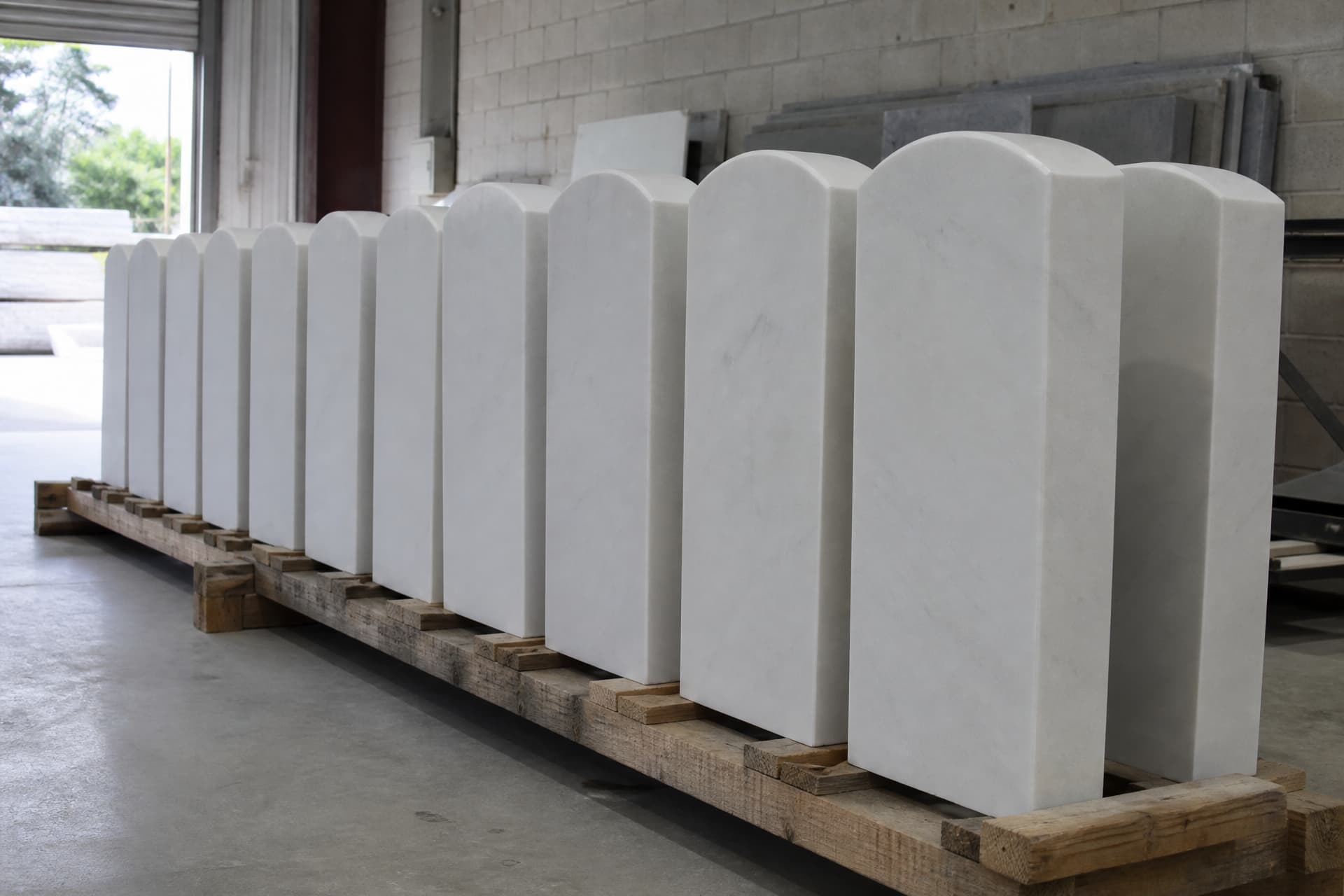 A row of new uncarved white marble government headstone blanks standing on a wooden rack in a monument works yard, every one identical in size and shape and entirely unlettered