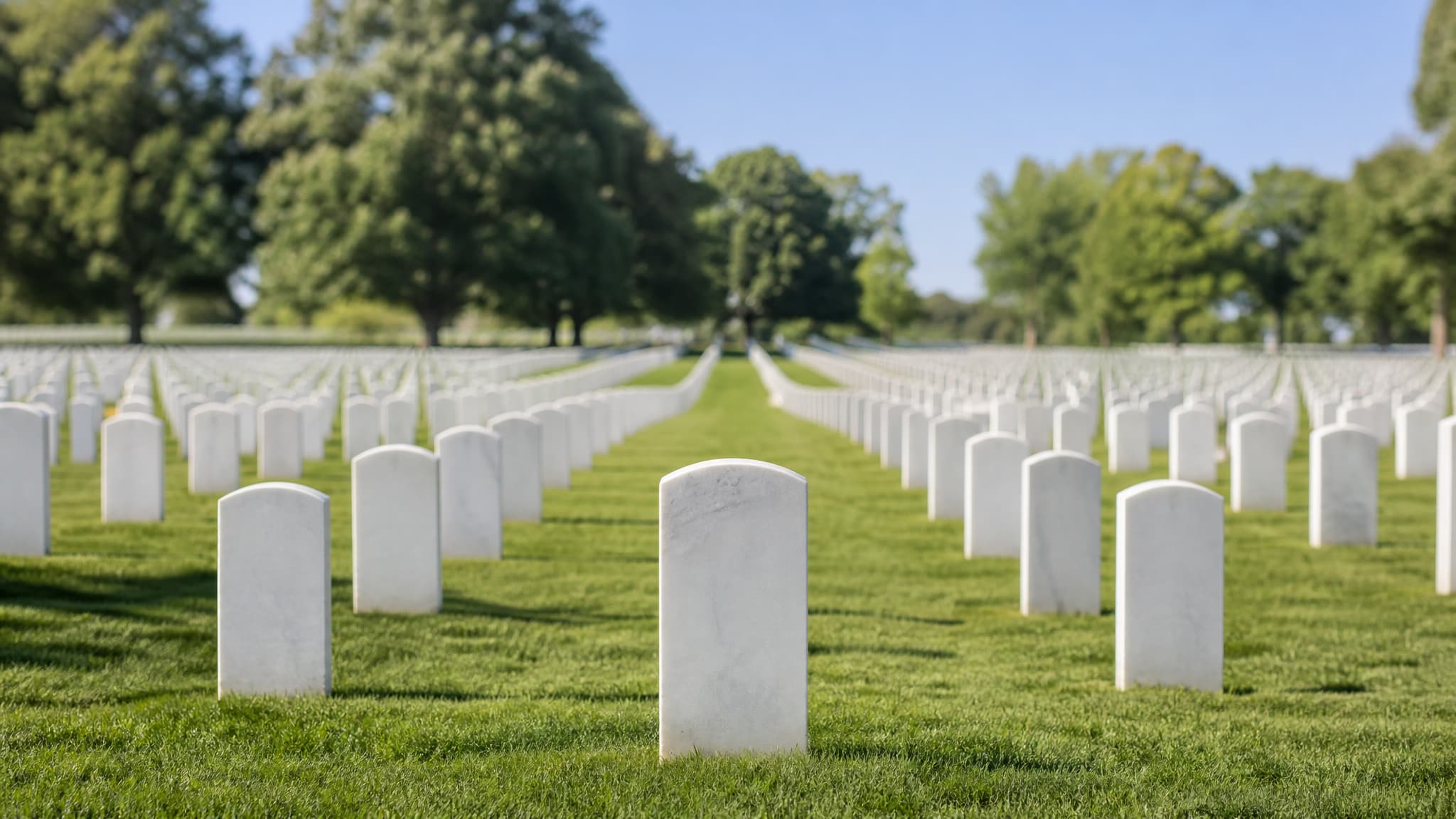 Rows of identical white marble headstones receding across mown green lawn on a gentle hillside under a clear sky, with mature trees along the ridge behind