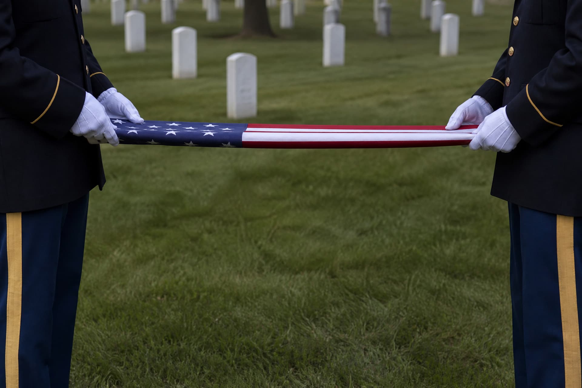 Two soldiers in dark dress uniform and white gloves holding a United States flag stretched taut between them at a graveside, mid-fold, seen from chest height downward with no faces in frame