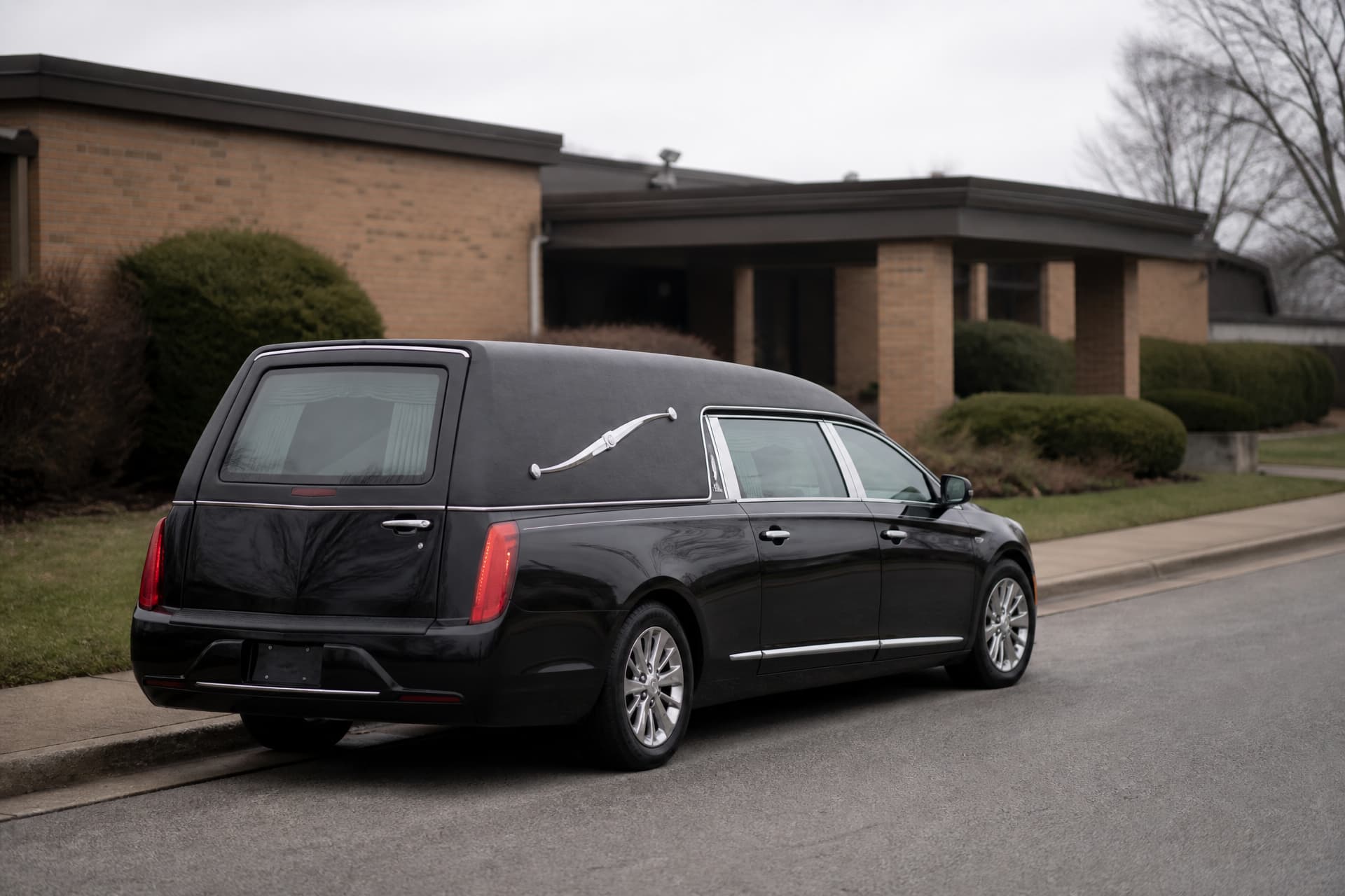 A black hearse parked outside a low brick funeral home on a quiet residential street in flat overcast daylight, rear door closed and nobody about