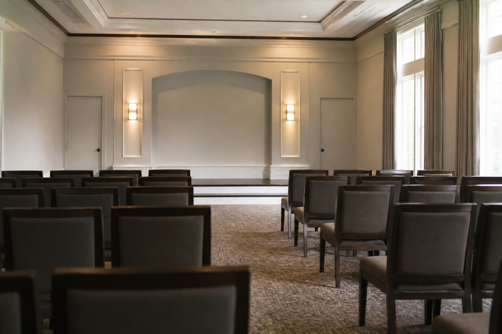 An empty funeral home chapel seen from the back row, rows of wooden chairs facing a low platform, tall windows down one side, no flowers and no casket
