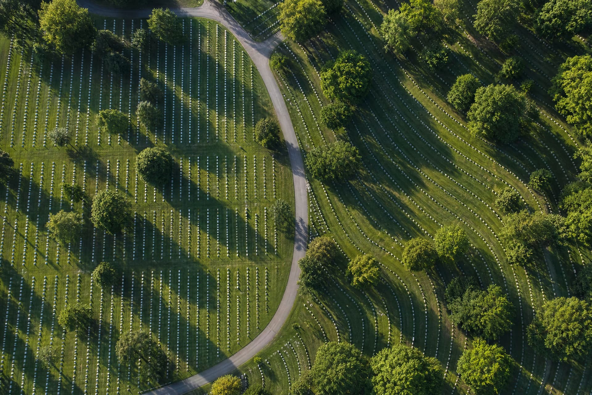 An aerial view looking straight down at a cemetery in morning light where two sections meet along a curving road, the older section laid out in rigid ranks squared to the compass and the newer one bending to follow the road and the slope