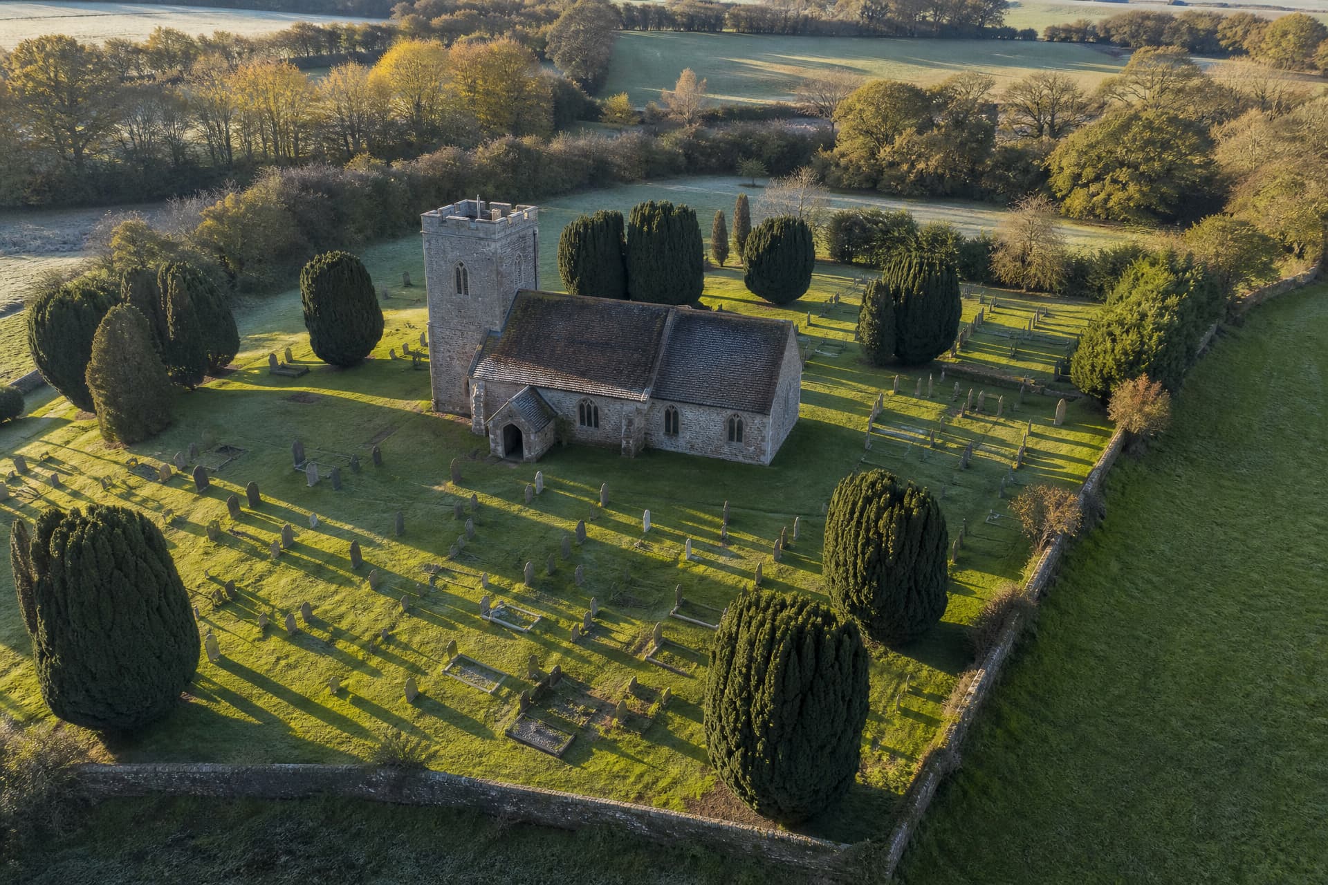 A low aerial view of an English parish churchyard on a clear morning, the stone church with its square tower lying on an east to west axis and the ranks of graves around it all running the same way