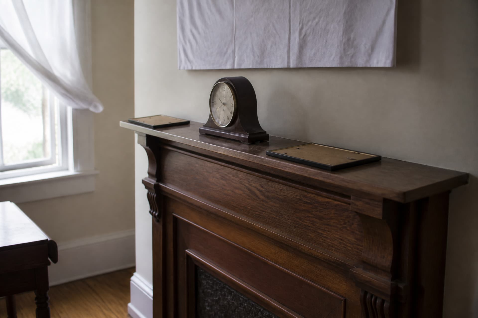 A mantel seen at a three-quarter angle with a stopped clock, two picture frames laid face-down on the wood, and the edge of a draped mirror above, an open window behind