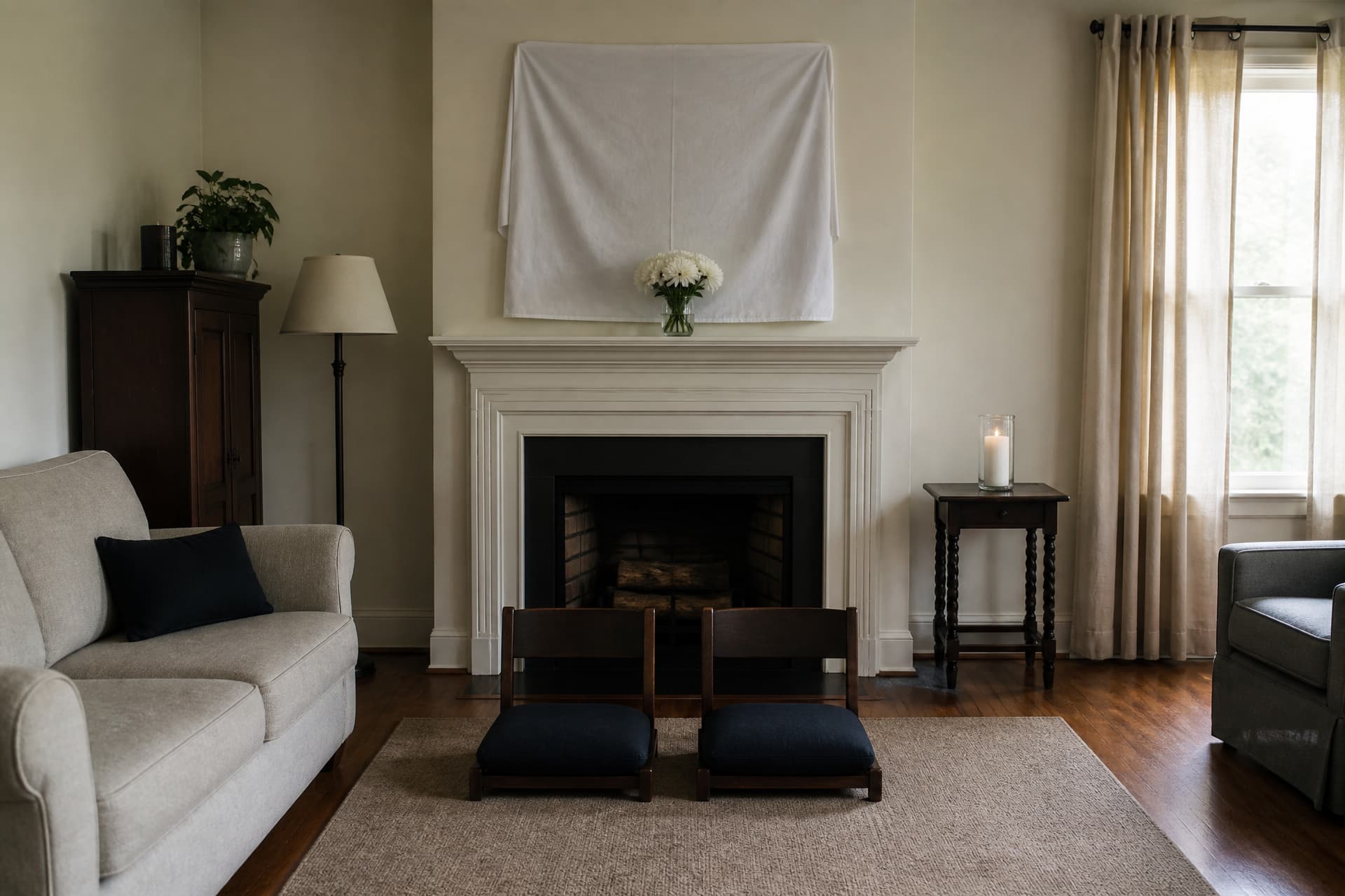 A modest living room set for a house of mourning with two low wooden chairs on the rug, a mirror above the mantel fully covered with a white cloth, and a single memorial candle burning in a glass on the side table