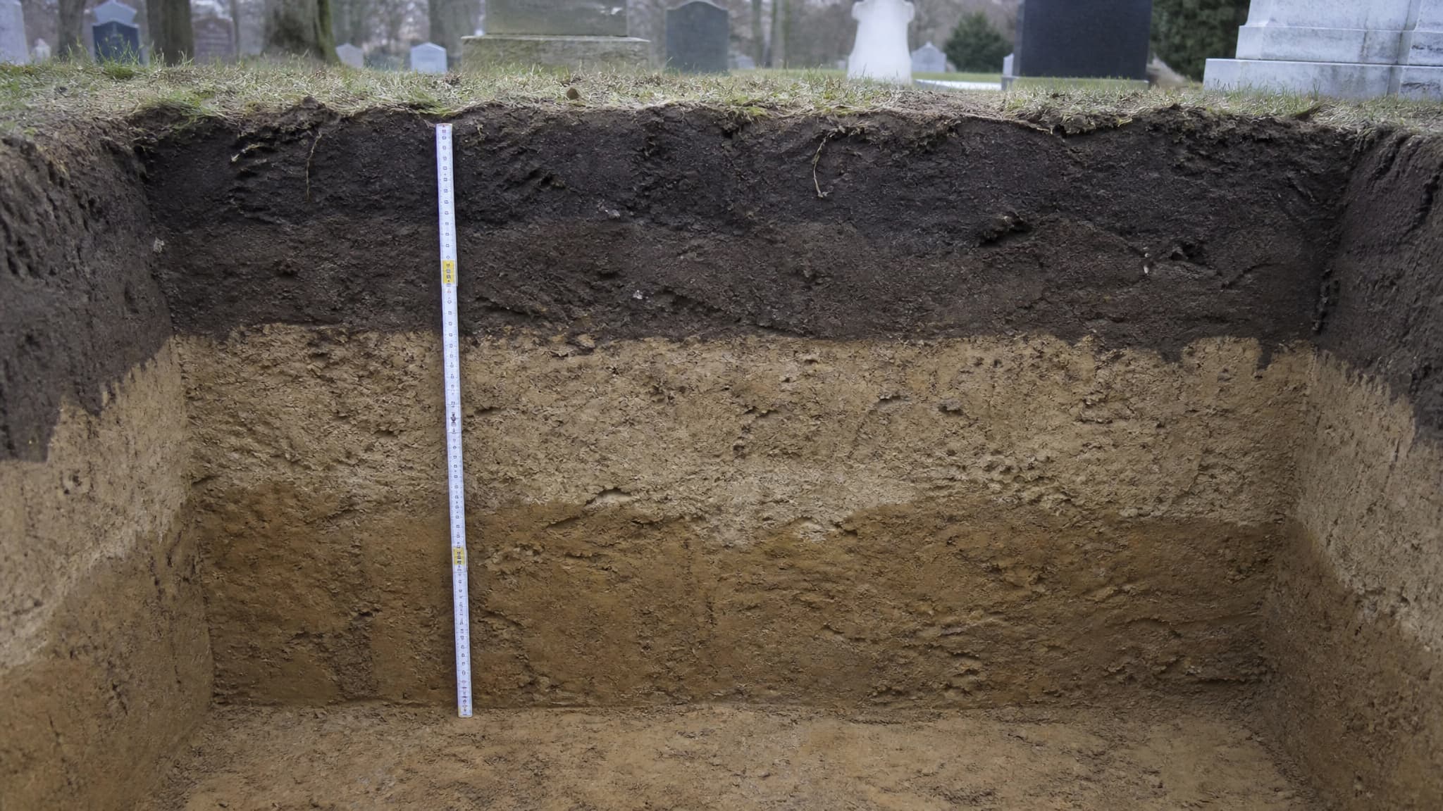 A cutaway diagram-like view of an open grave in cemetery ground seen from the side, showing the layered soil profile, a concrete burial vault set at the bottom and the measured depth of earth cover above it, in flat overcast daylight