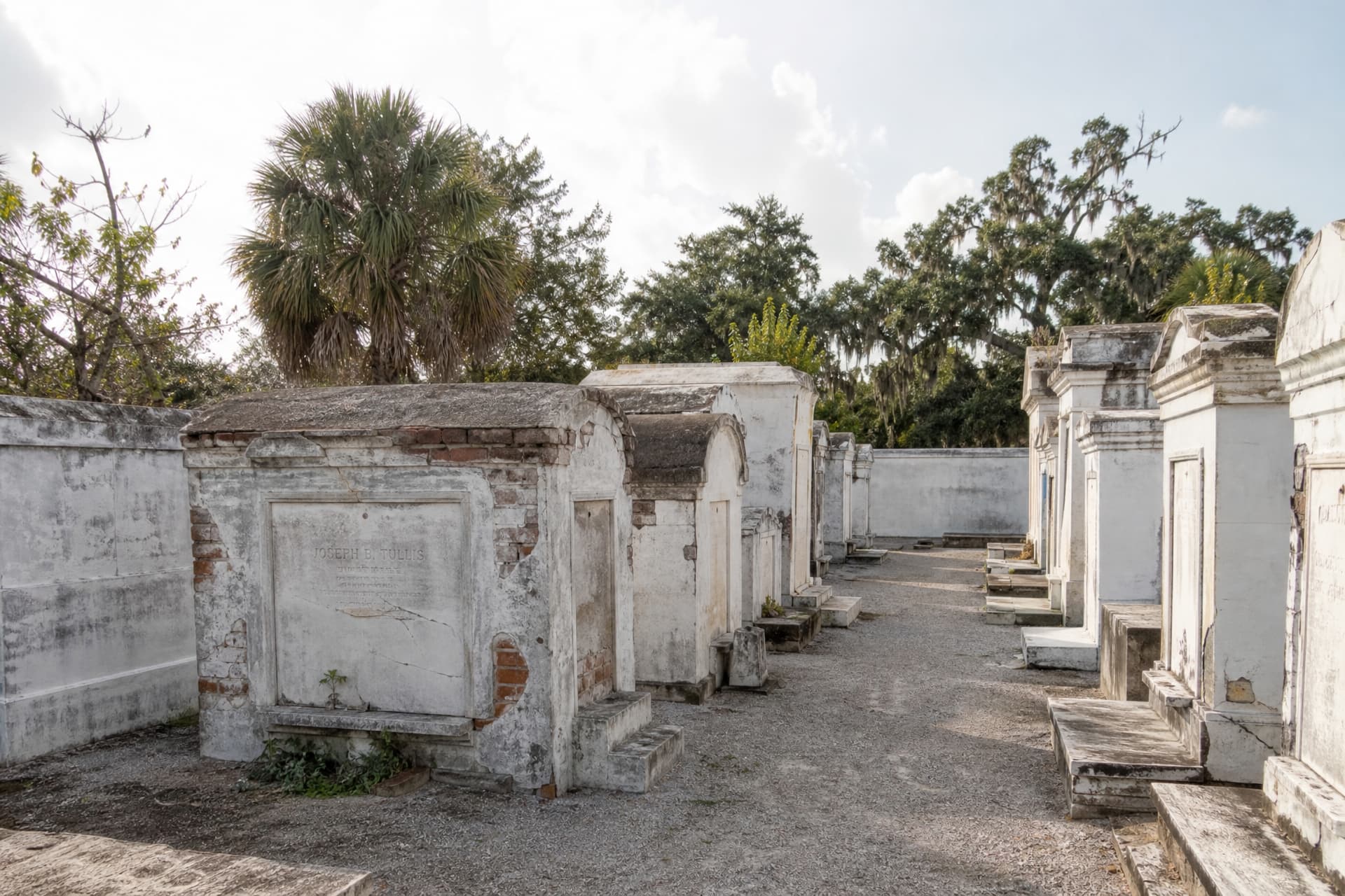 A row of whitewashed above-ground brick and stucco tombs in a southern American cemetery under a bright hazy sky, the vaults weathered and slightly leaning with narrow gravel paths between them