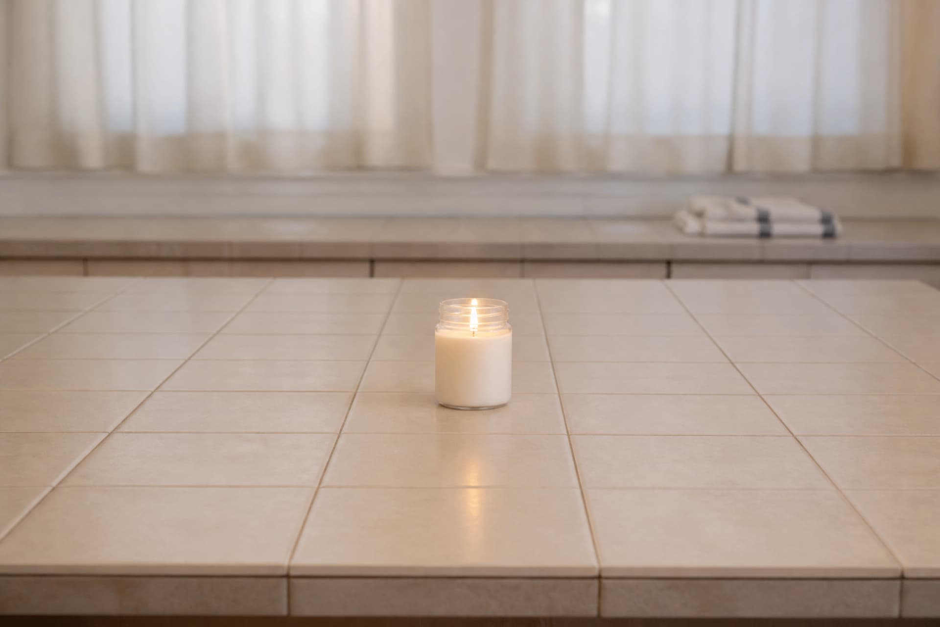 A jar candle burning alone on a tiled surface with wide empty space around it and curtains far in the background
