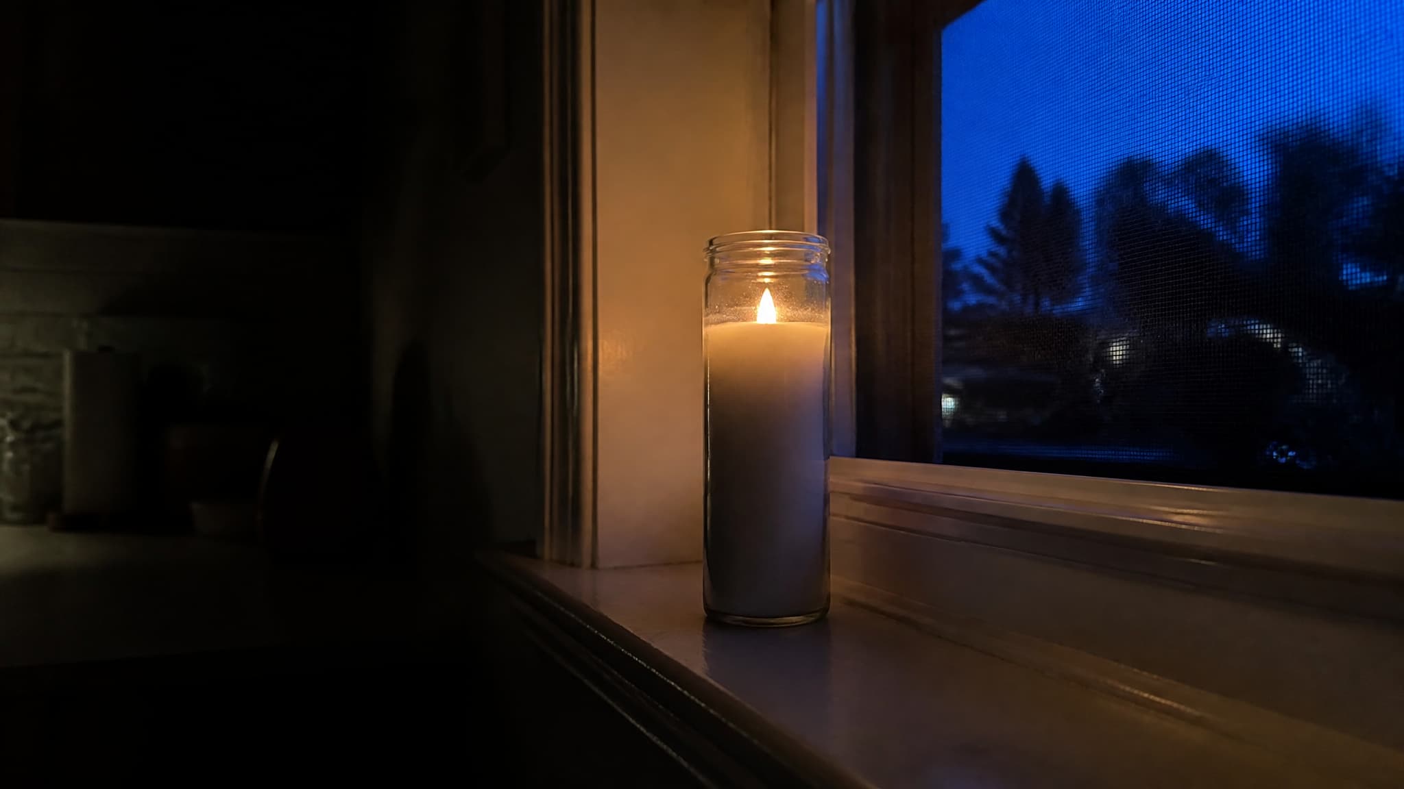 A single memorial candle in a plain glass jar burning on a kitchen windowsill against a deep blue post-sunset sky